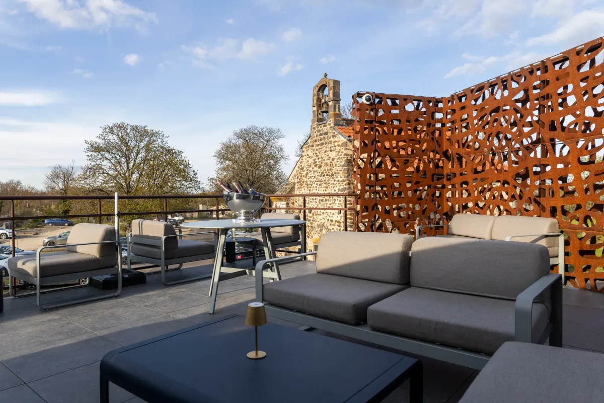 Terrasse avec canapés, table et lampe, vue sur une église en pierre et un parking