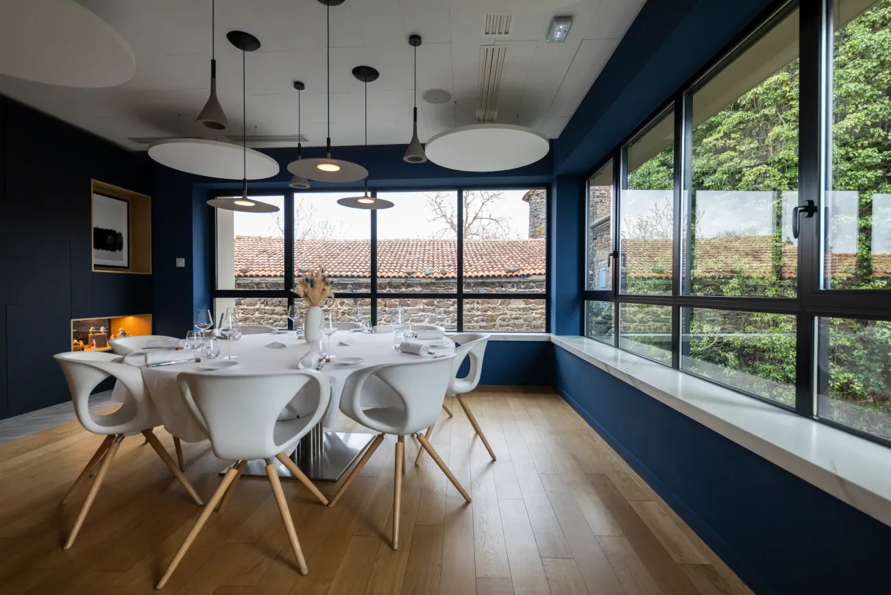 Salle à manger moderne avec table blanche et chaises assorties, vue sur des toits en tuiles et végétation à travers de grandes baies vitrées
