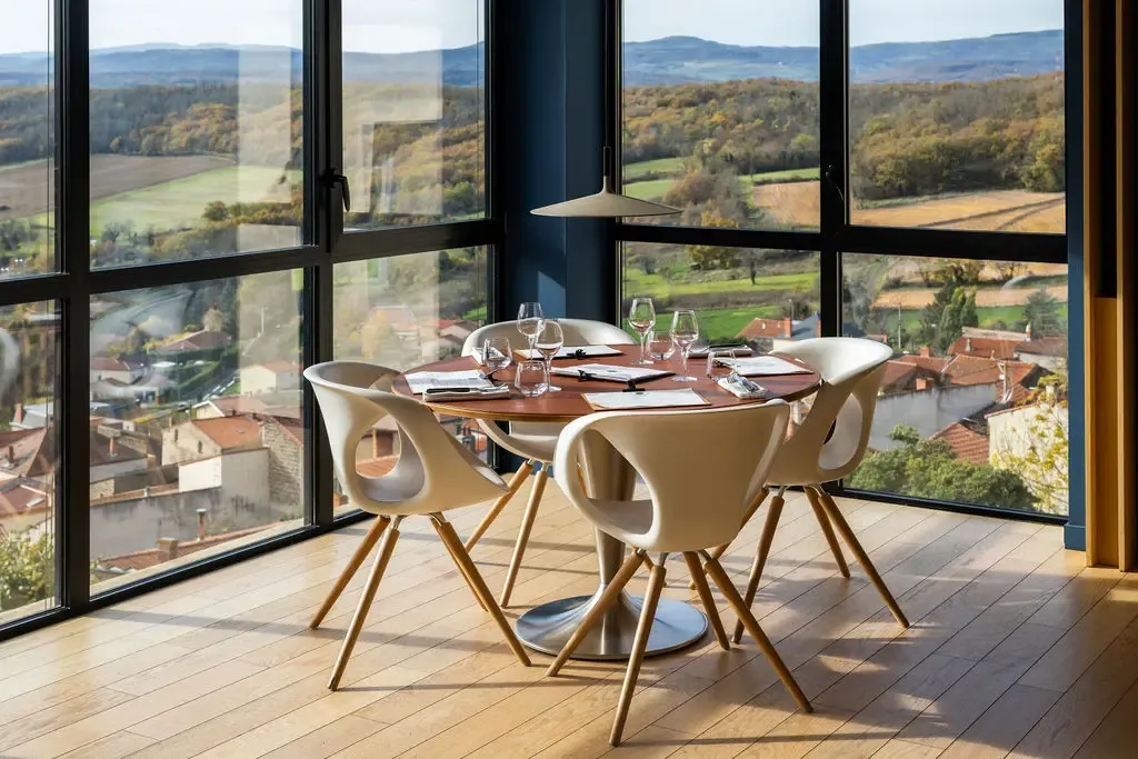 Round table with four modern chairs, glasses and cutlery, in front of large windows offering a view of a village and lush green hills.