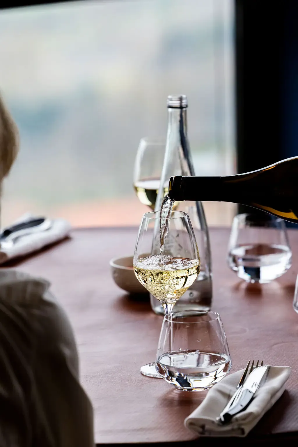 Une bouteille de vin blanc est versée dans un verre à pied sur une table avec des couverts et un verre d'eau