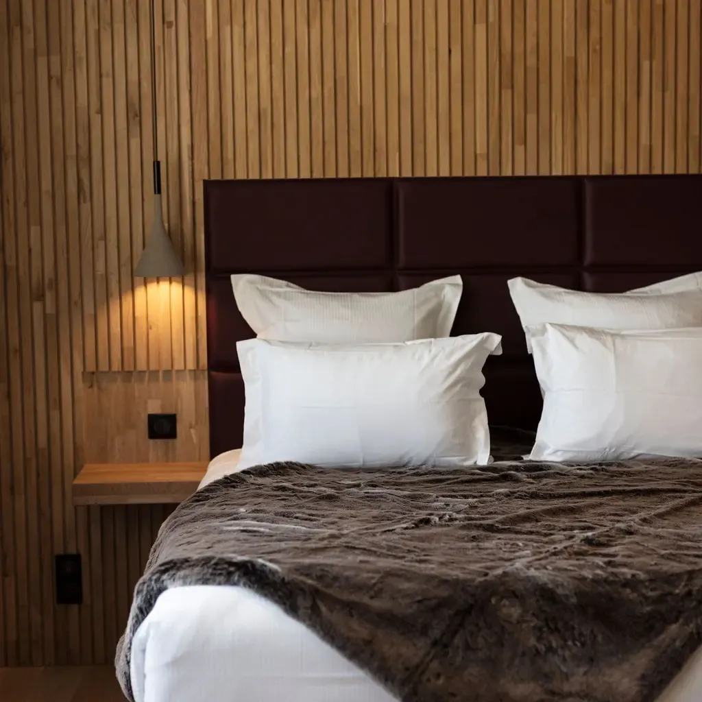 Lit avec une tête de lit en cuir marron, des draps blancs et une couverture grise dans une chambre aux murs en bois