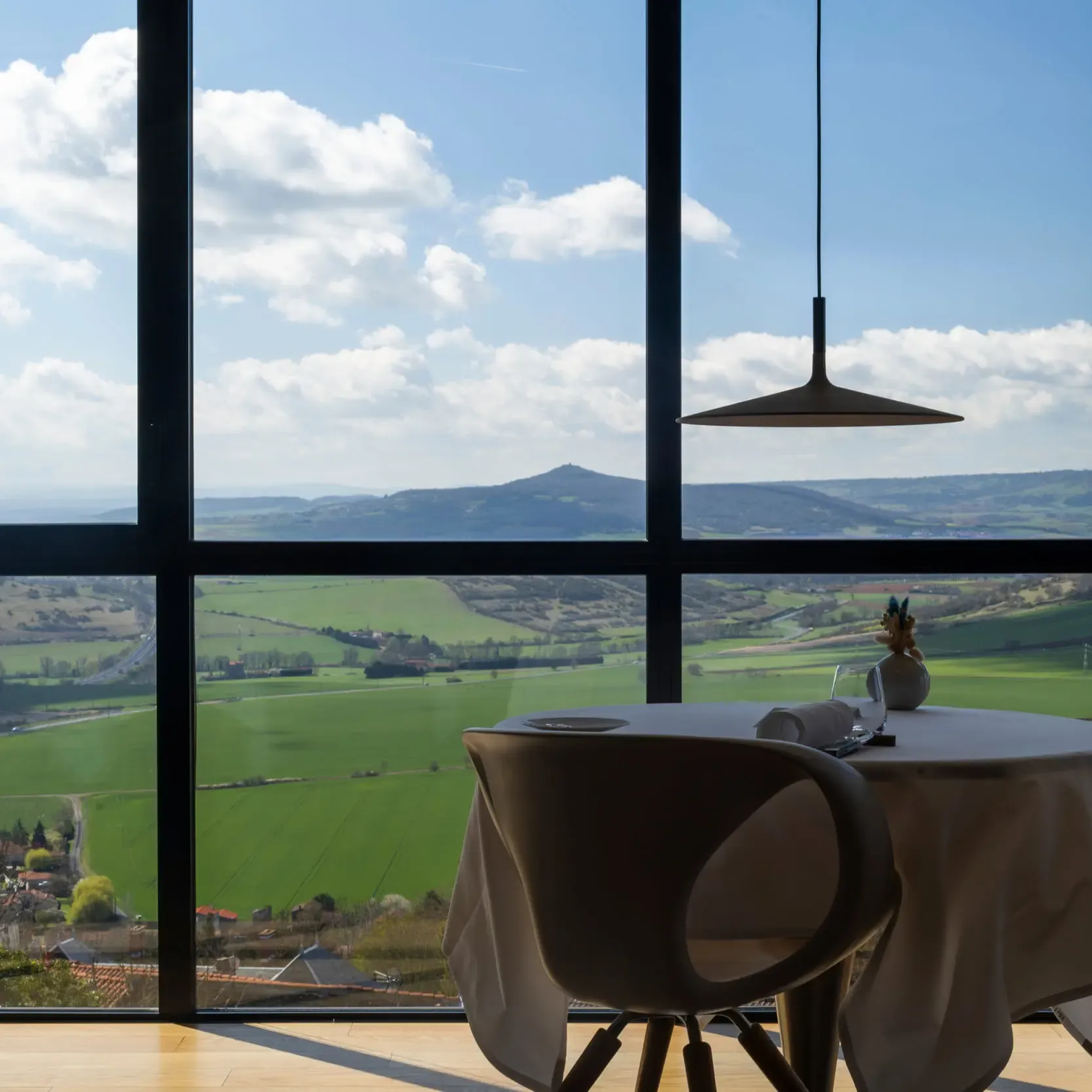 Une table avec une nappe et un vase de fleurs devant une grande fenêtre offrant une vue sur des collines verdoyantes et un ciel partiellement nuageux