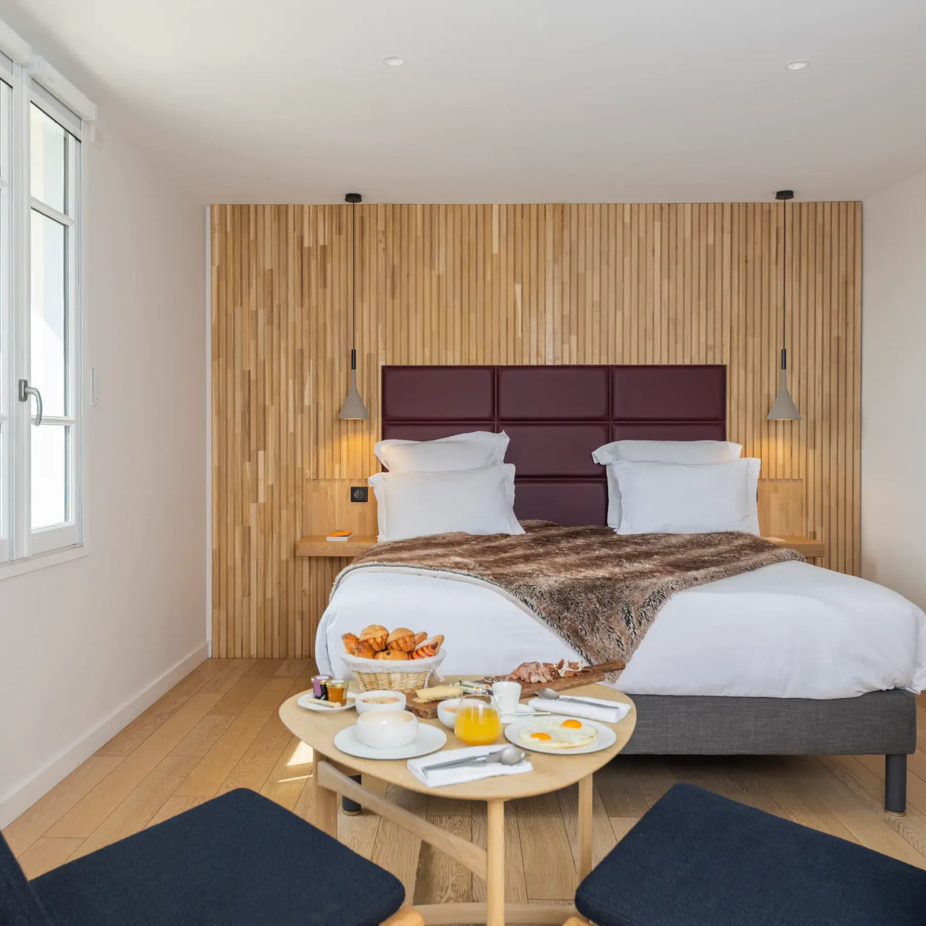Chambre avec lit double, tête de lit en bois et velours, table avec petit-déjeuner, deux chaises et fenêtre