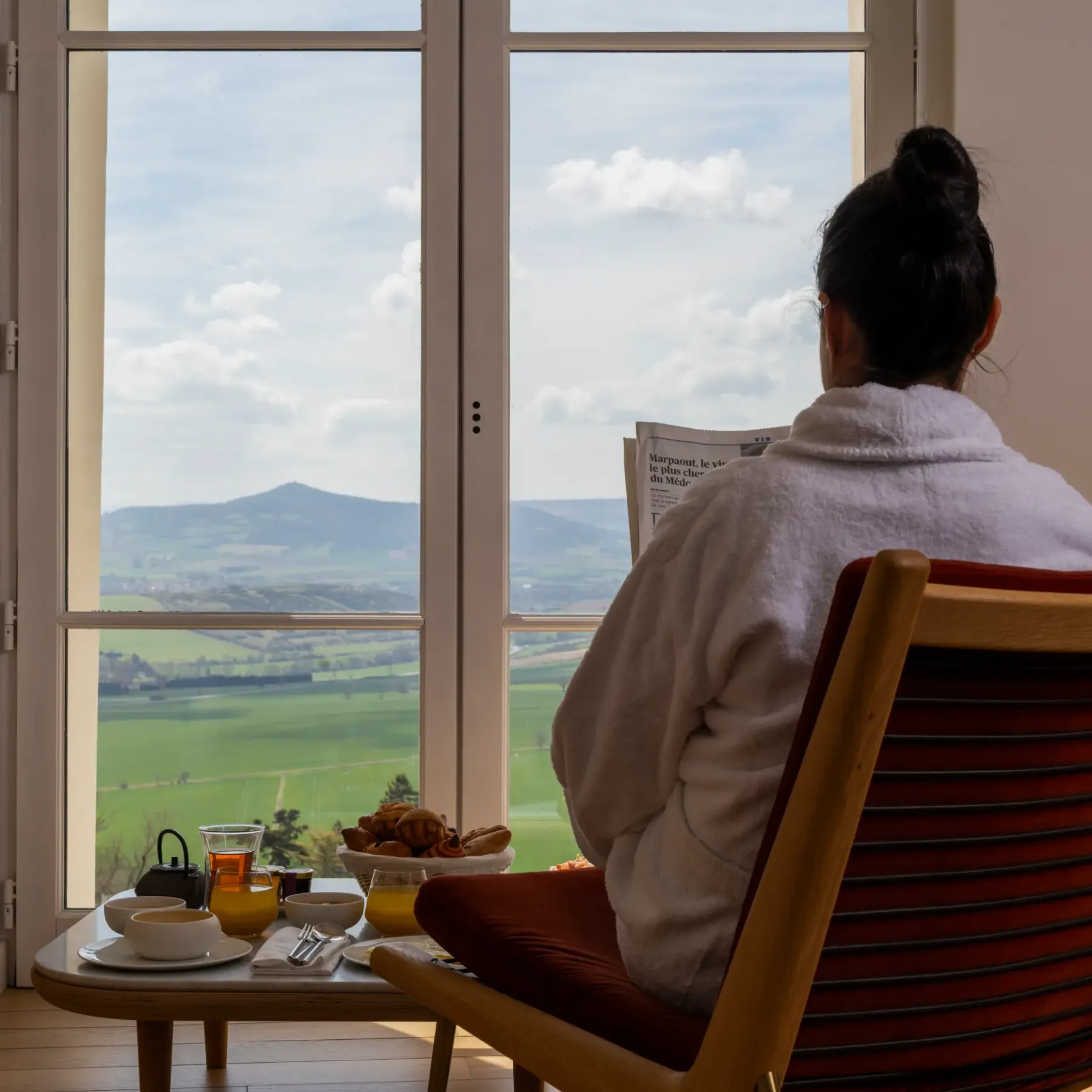 Une personne assise dans un fauteuil face à une fenêtre ouverte, avec une vue sur des collines verdoyantes, un plateau de petit-déjeuner à côté d'elle