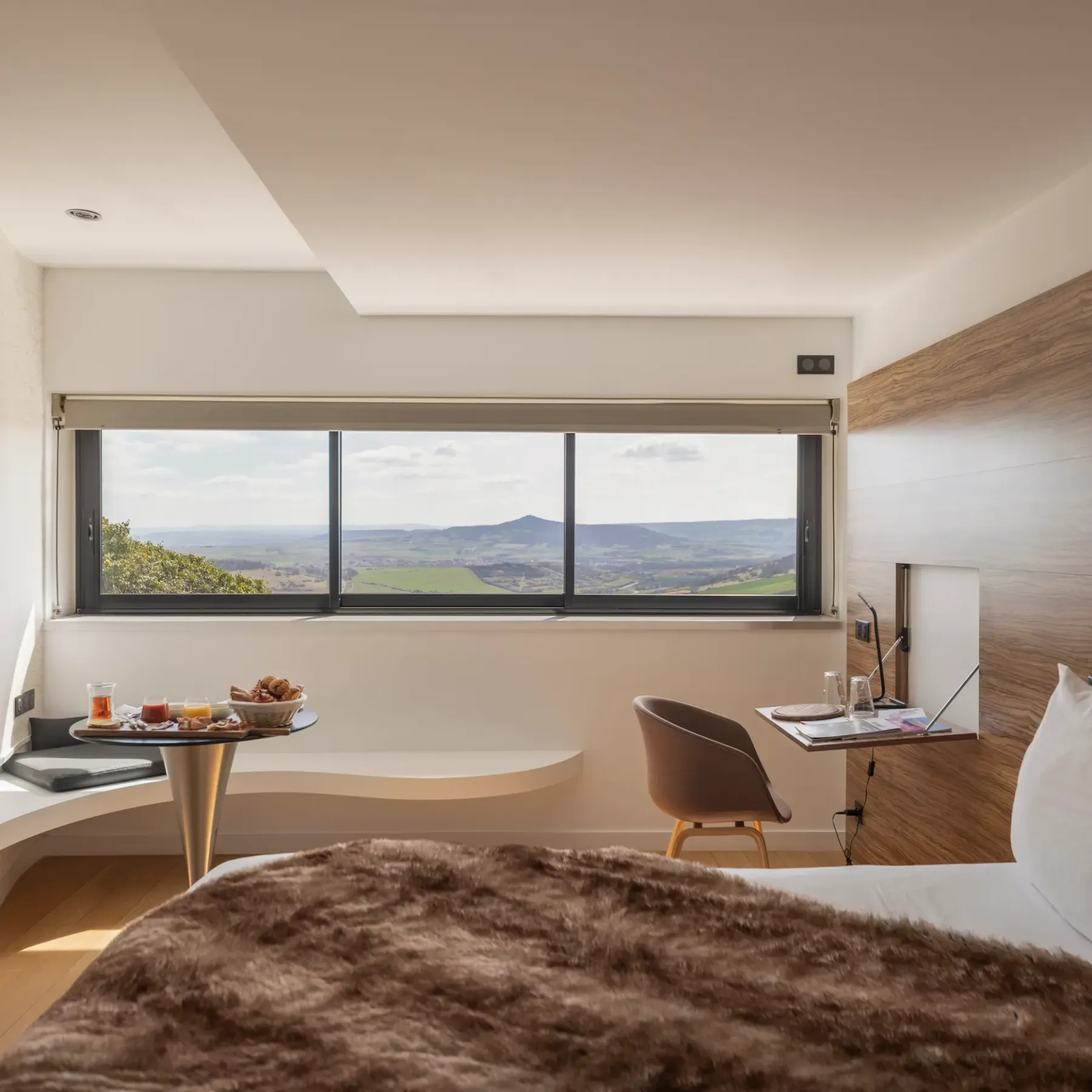 Chambre avec lit, table de chevet, chaise et bureau, offrant une vue panoramique sur un paysage vallonné à travers une grande fenêtre