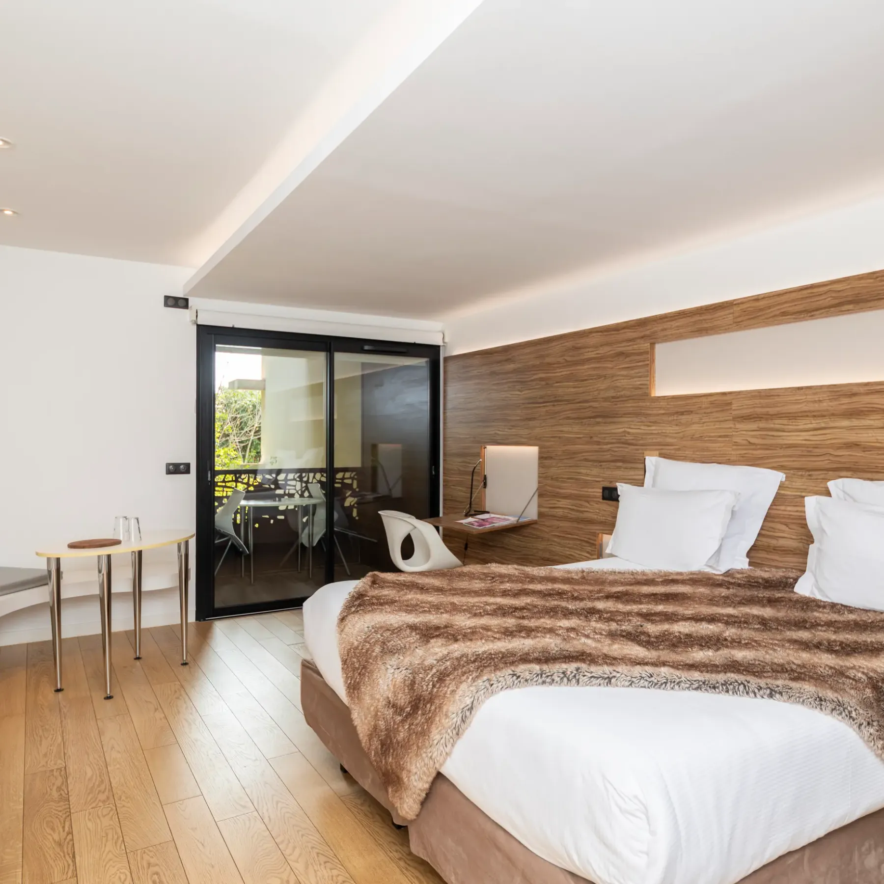 Chambre moderne avec un lit double, une table de chevet, une porte-fenêtre donnant sur un balcon et un mur en bois