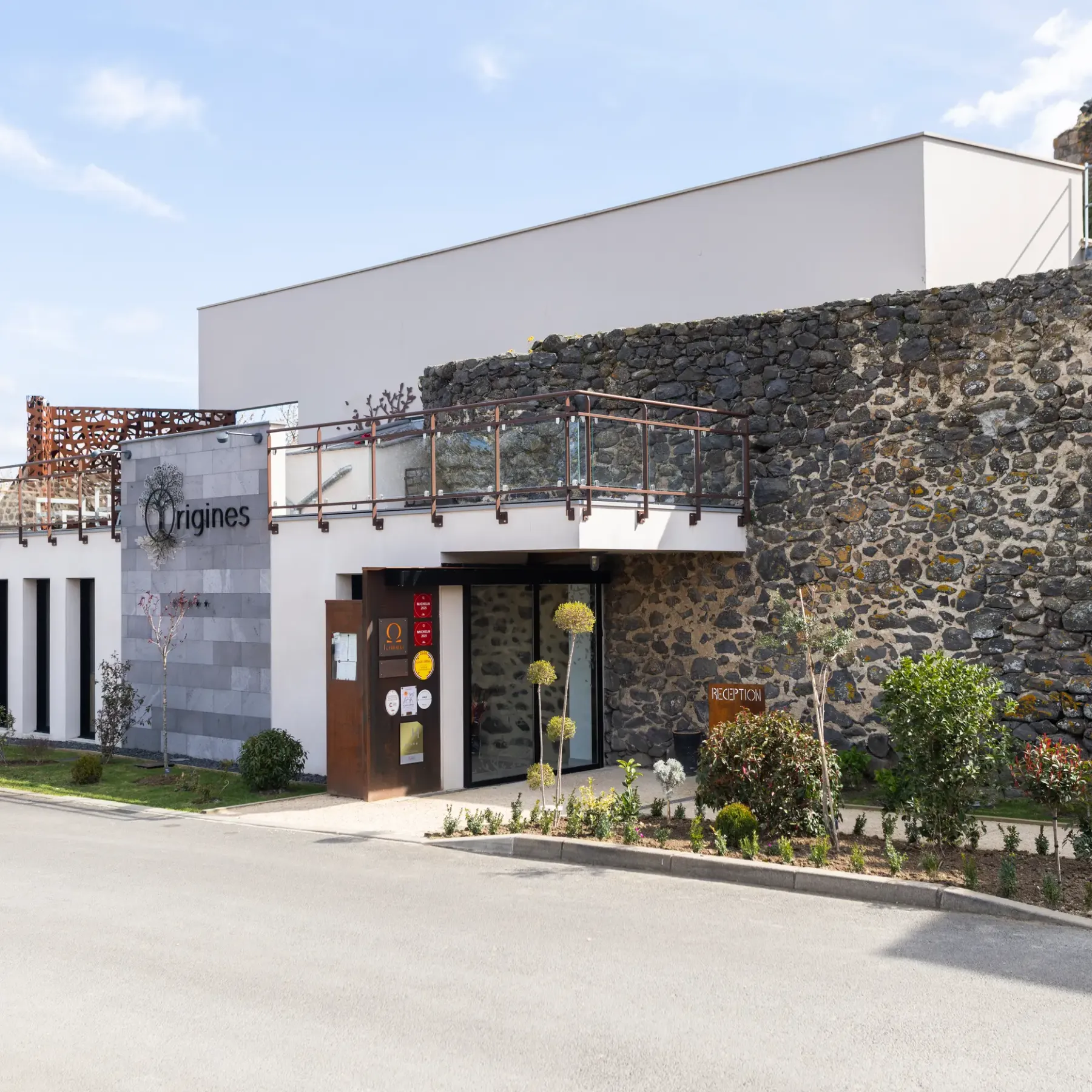 Bâtiment moderne avec façade en pierre et balcons, portant l'enseigne "Origines" et situé dans un cadre paysager