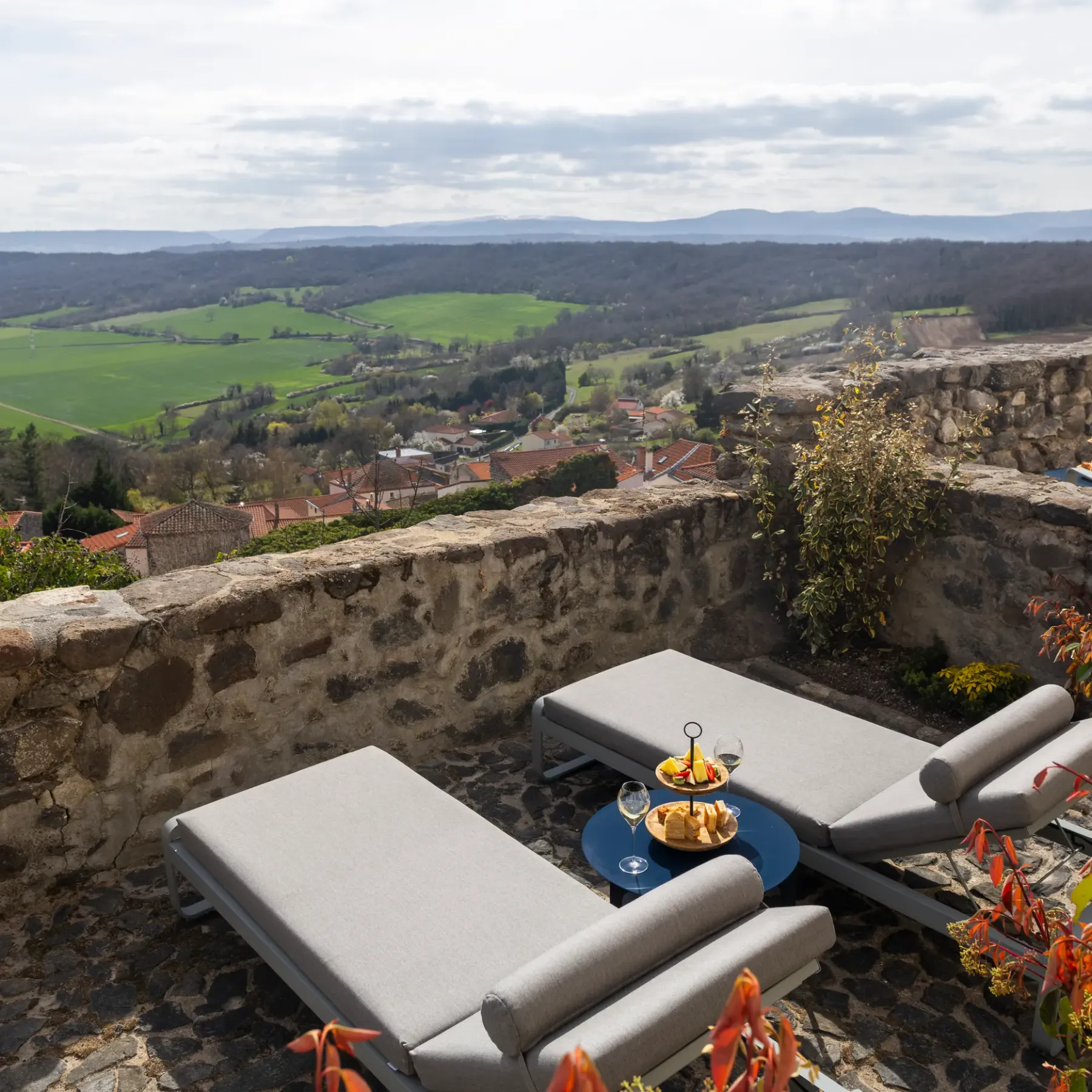 Deux chaises longues grises avec une table basse sont disposées sur une terrasse en pierre offrant une vue sur une vallée verdoyante et des collines lointaines
