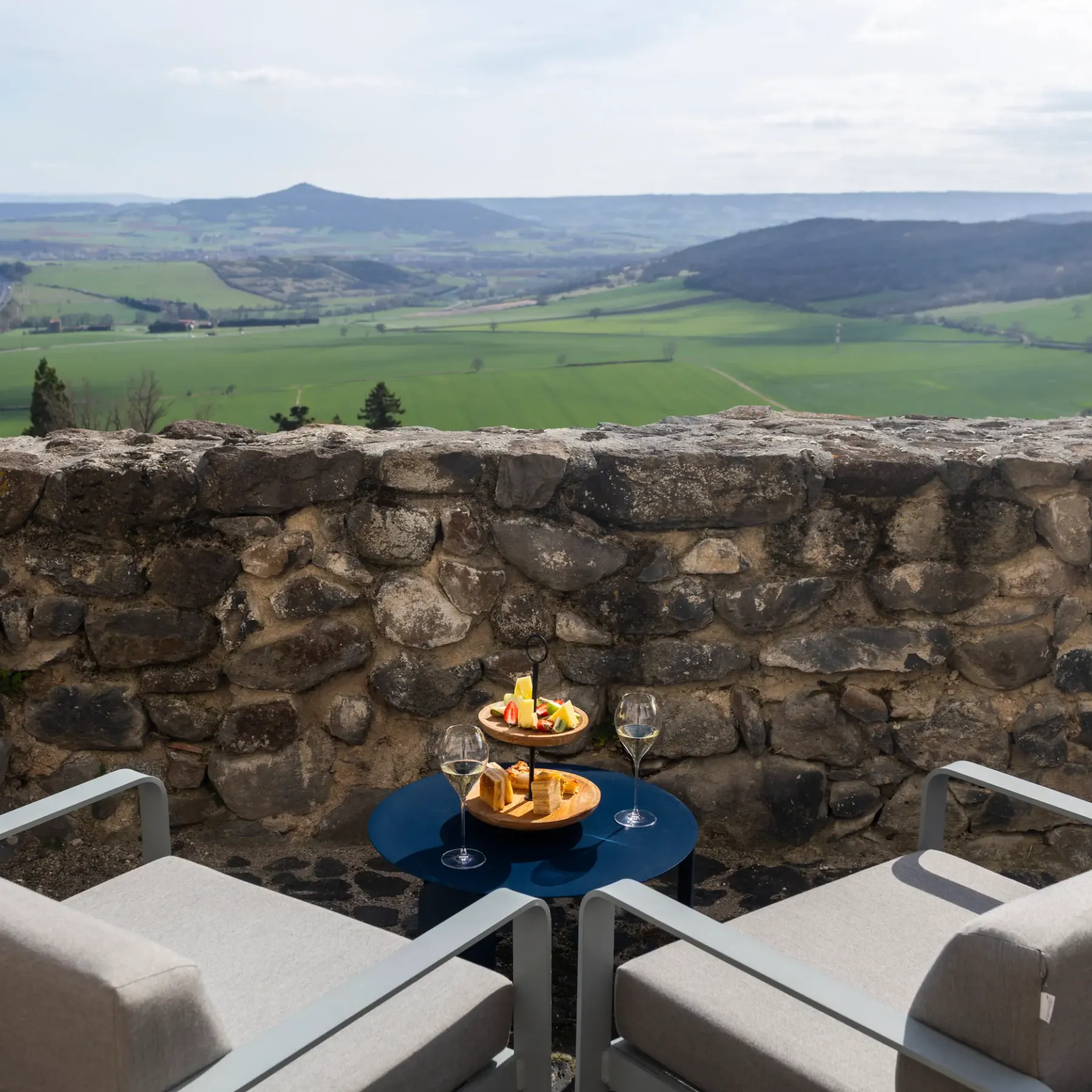 Une table avec des verres et une assiette de nourriture est installée devant un mur de pierre, offrant une vue sur une vallée verdoyante