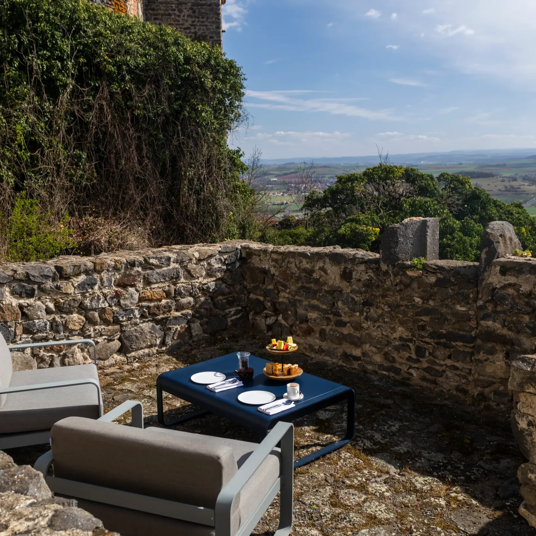 Un espace extérieur avec deux chaises longues, une table basse et un plateau de petit-déjeuner, entouré de murs de pierre et offrant une vue sur un paysage vallonné