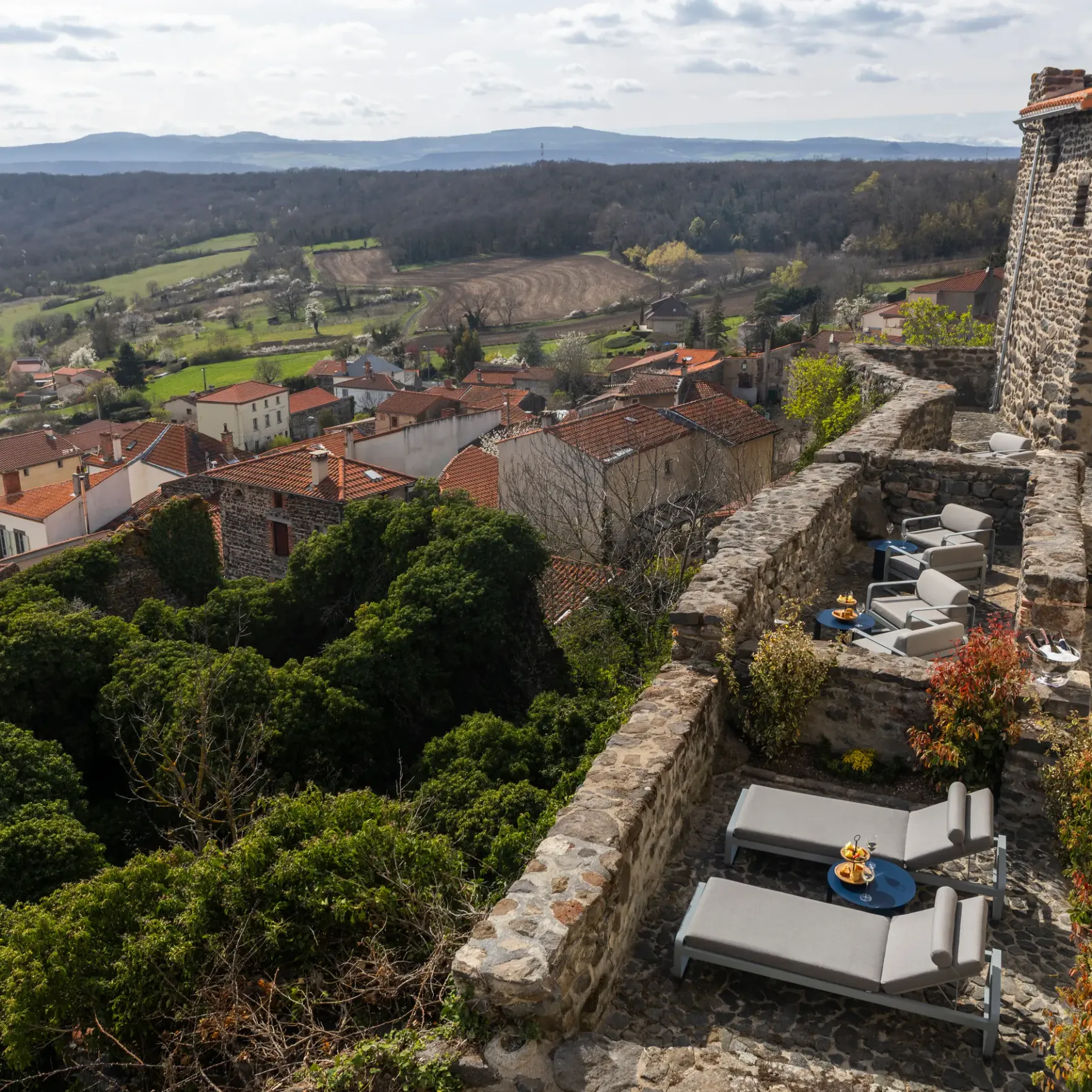 Vue d'un village avec des toits rouges, entourée de verdure et de champs, depuis une terrasse avec des chaises longues et une table