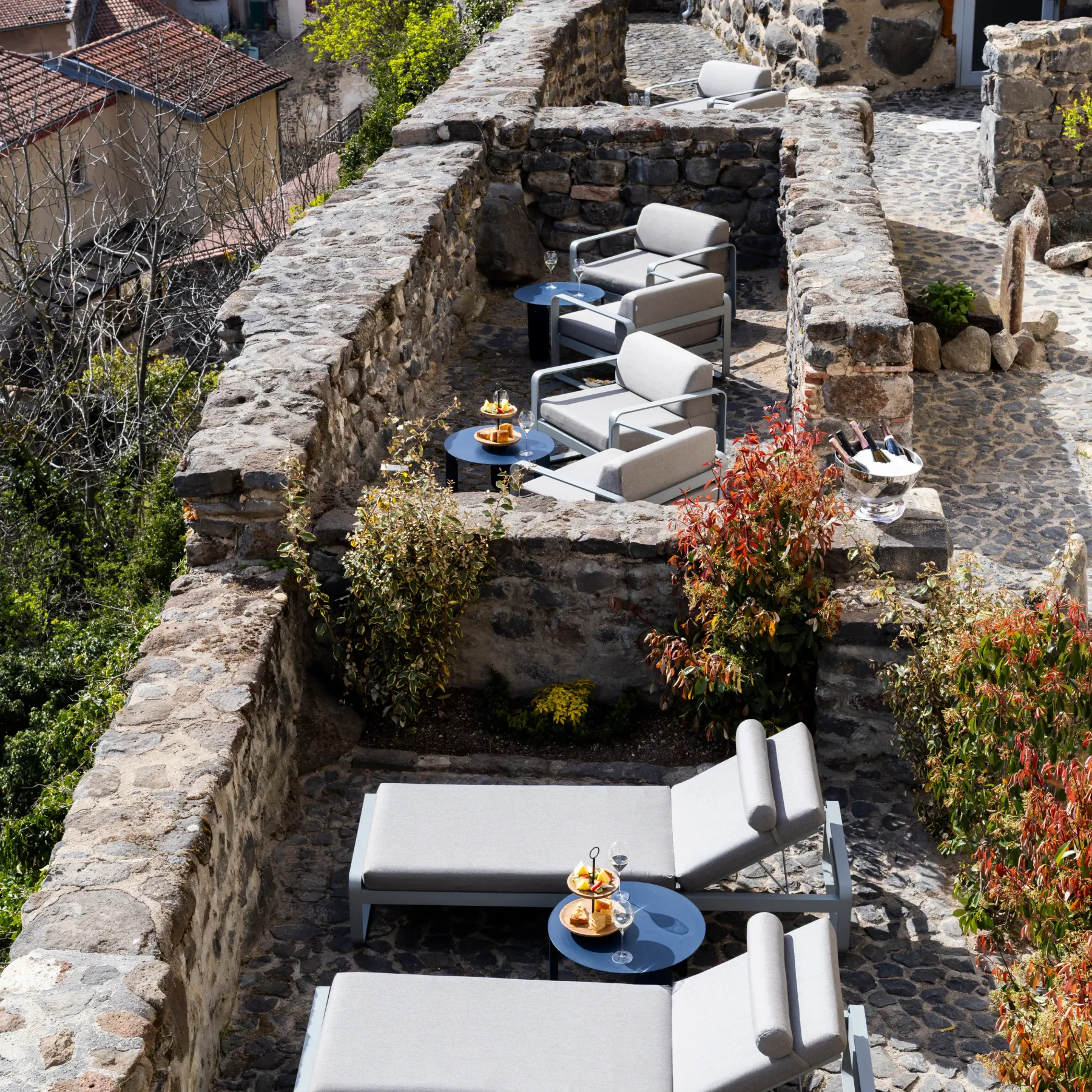 Terrasse en pierre avec des chaises longues et des tables basses, surplombant un village aux toits de tuiles rouges et aux murs de pierre