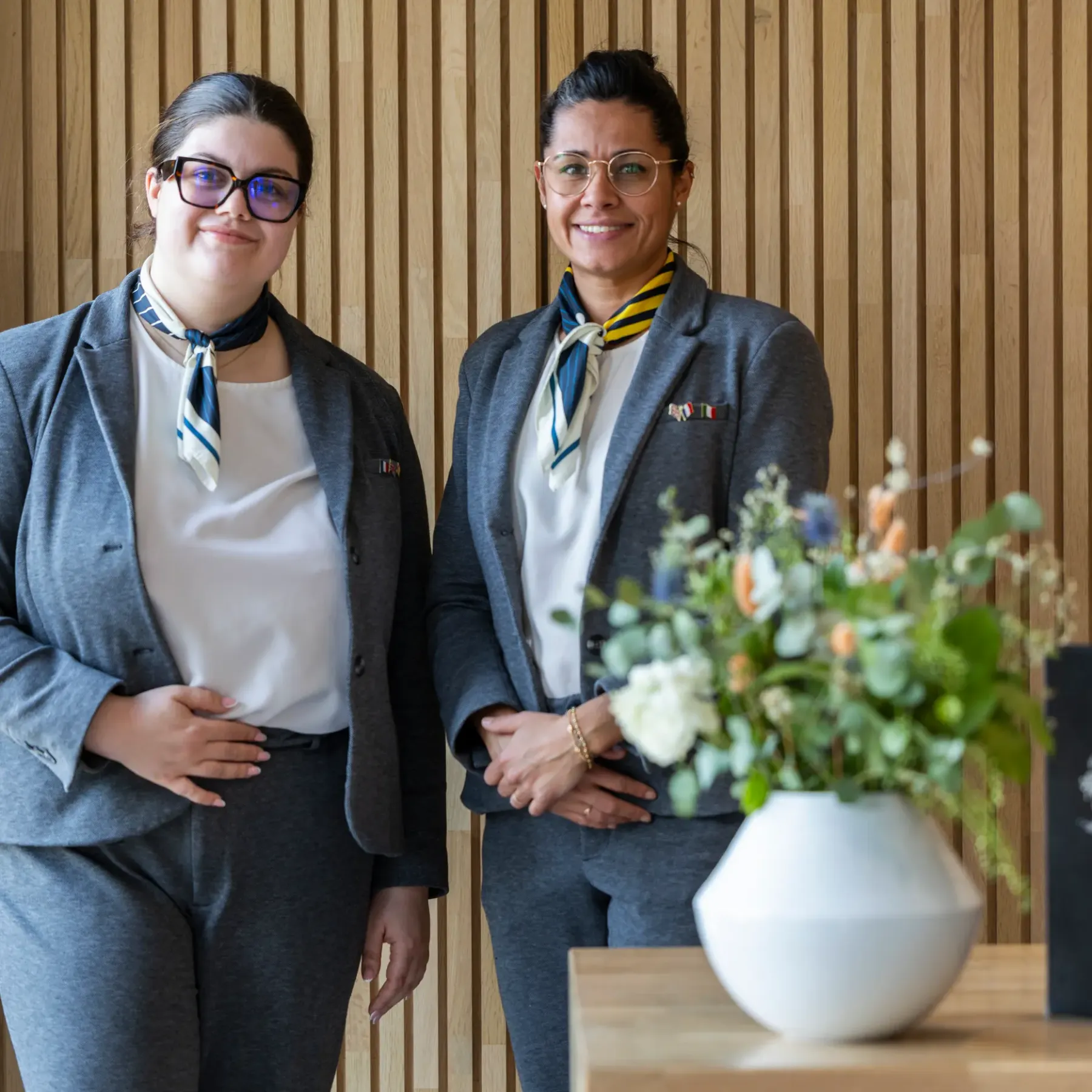 Deux femmes en costume avec des foulards et des badges se tiennent devant un mur en bois, avec un vase de fleurs et un ordinateur portable sur une table à côté d'elles