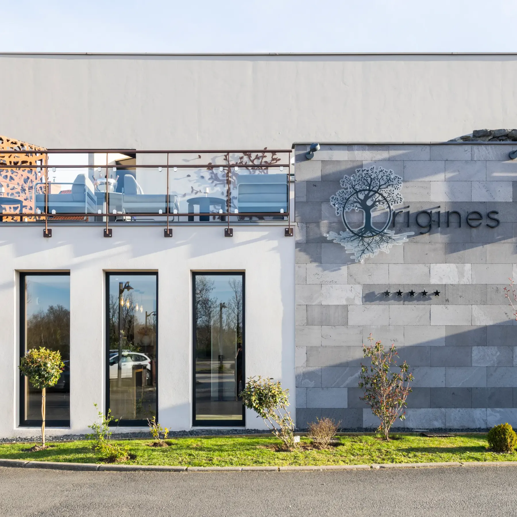 Bâtiment moderne avec façade blanche et grise, balcon à balustrade métallique et végétalisée, et enseigne "Origines" accompagnée d'un logo d'arbre
