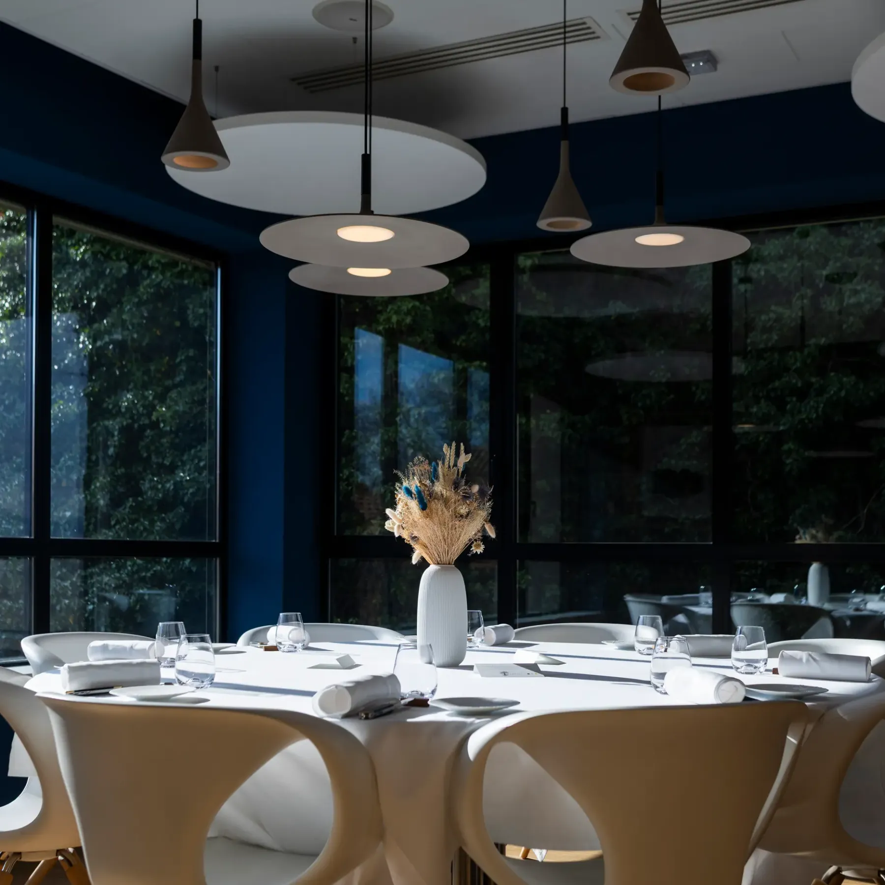 Table dressée avec des verres et des serviettes dans une salle à manger moderne aux grandes baies vitrées, éclairée par des suspensions circulaires