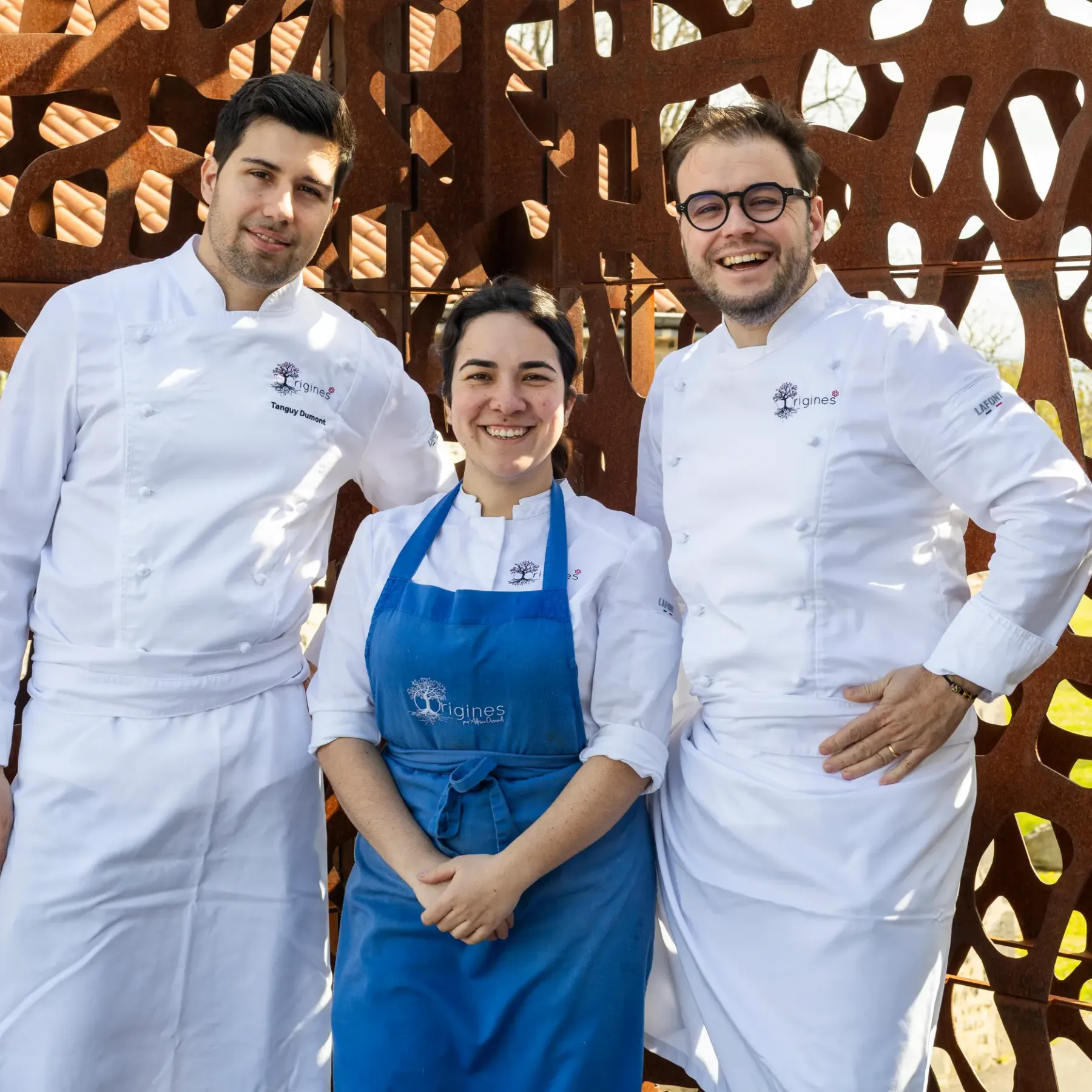 Trois personnes en tenue de cuisine professionnelle posent devant une structure métallique ajourée en extérieur
