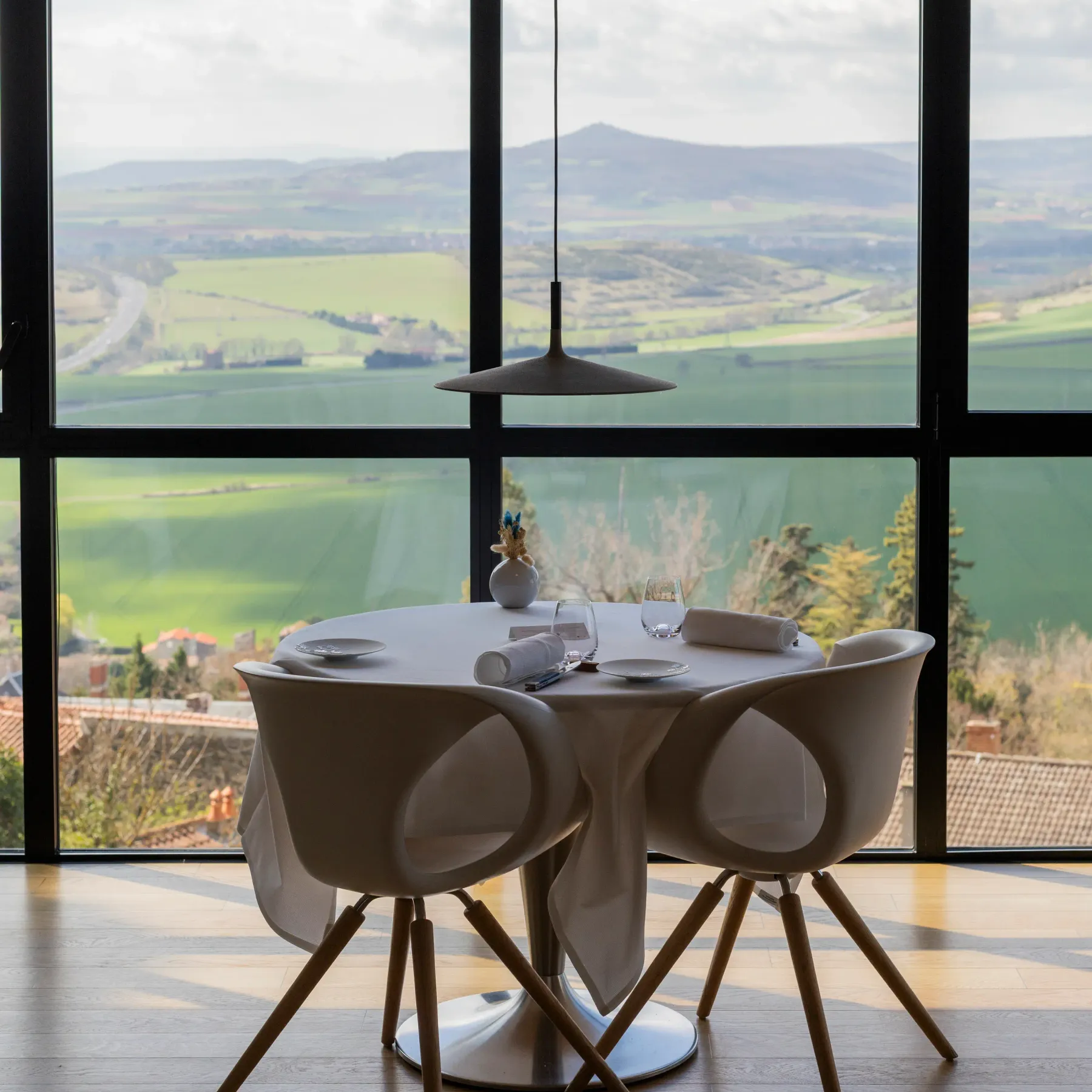 Table ronde avec deux chaises devant une grande baie vitrée offrant une vue sur un paysage vallonné