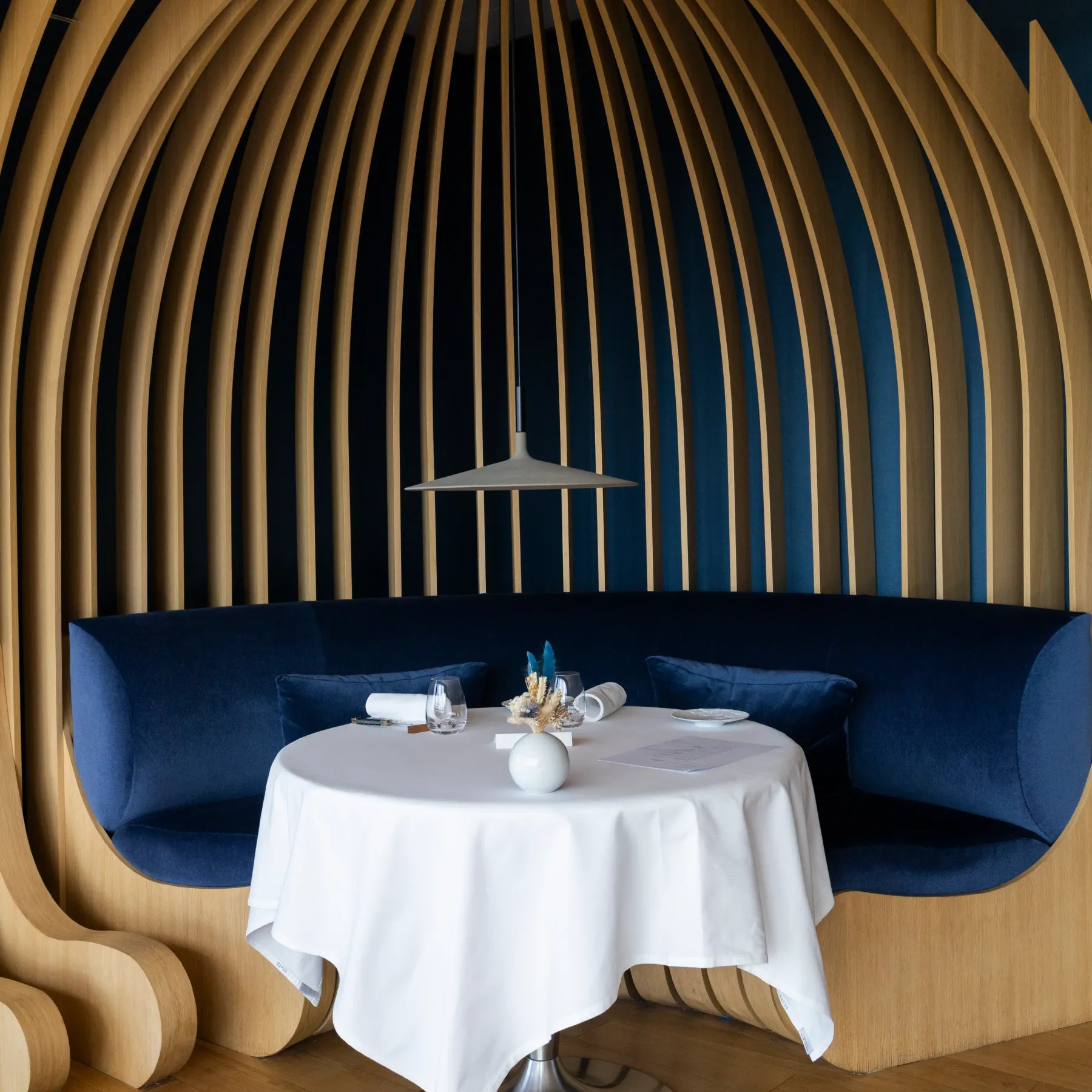 Un espace intérieur avec une table ronde recouverte d'une nappe blanche, entourée de banquettes bleues, sous une structure en bois et un éclairage suspendu