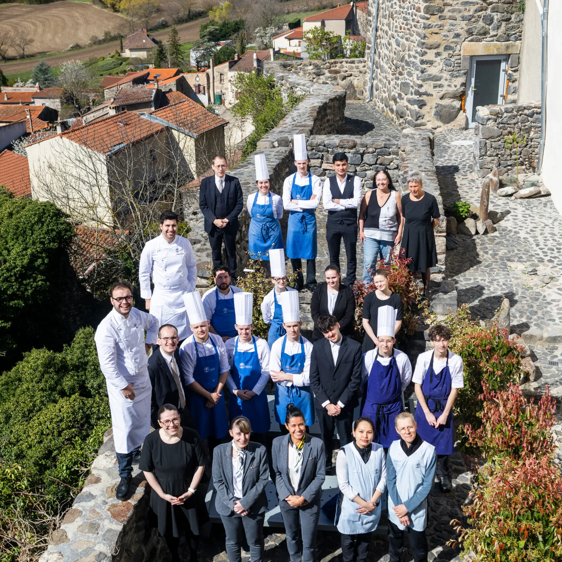 Un groupe de personnes en tenue de cuisine et en vêtements formels pose devant un bâtiment en pierre dans un village avec vue sur la campagne