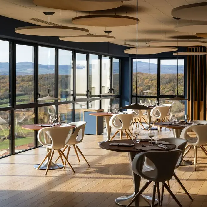 Modern restaurant with large windows offering a panoramic view of a hilly and wooded landscape, tables and chairs arranged on a light-colored parquet floor.