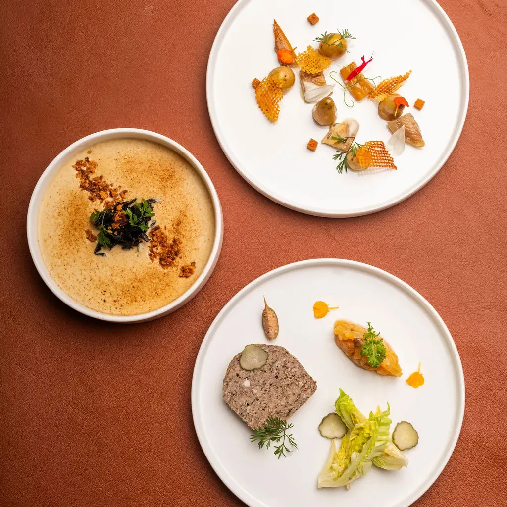 Three white plates featuring gourmet dishes arranged with precision against an orange background