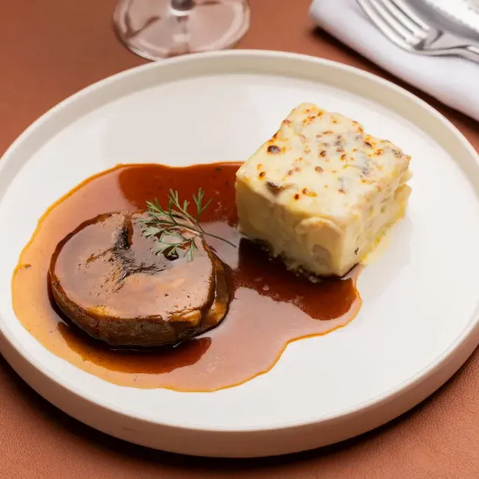 A piece of meat with a brown sauce and parsley garnish, served with a piece of gratin, all on a white plate.