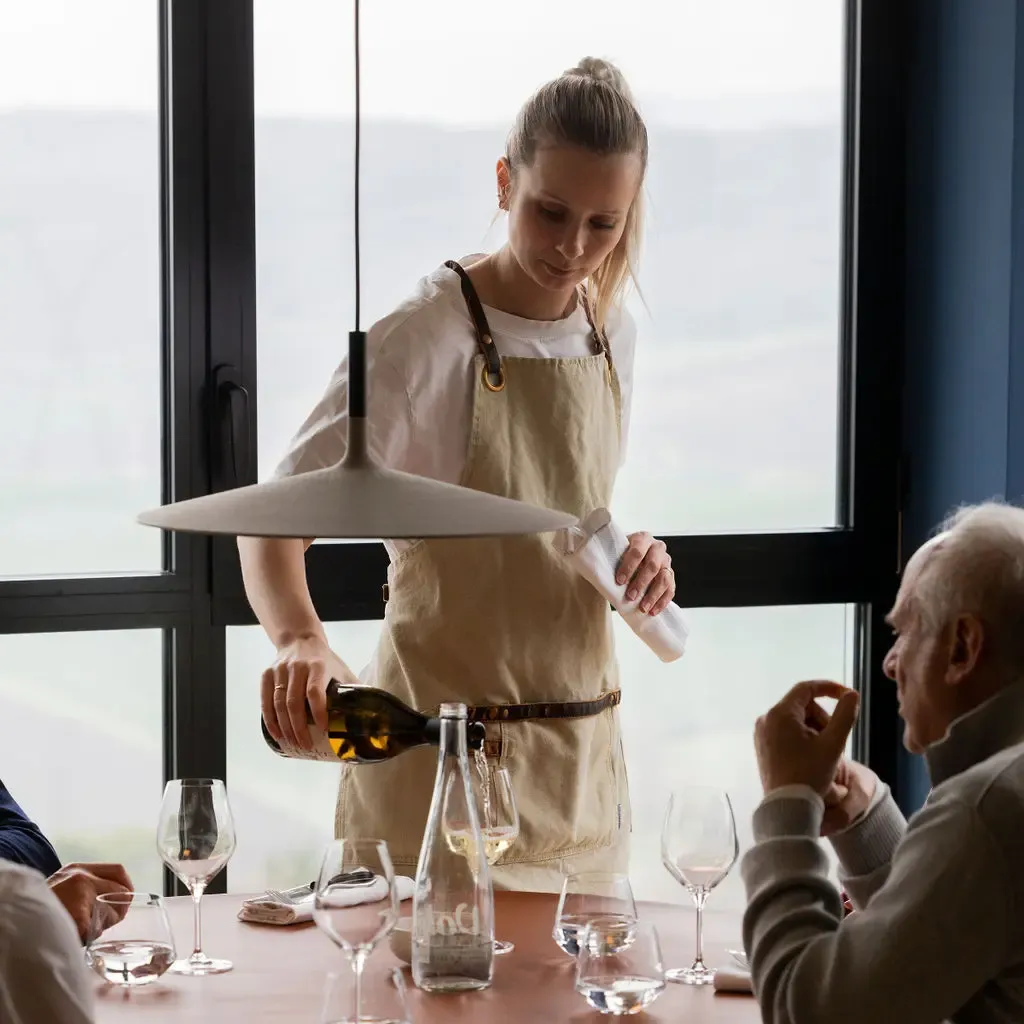 A waitress in a beige apron pours wine for a customer seated at a table with wine glasses and a water carafe.