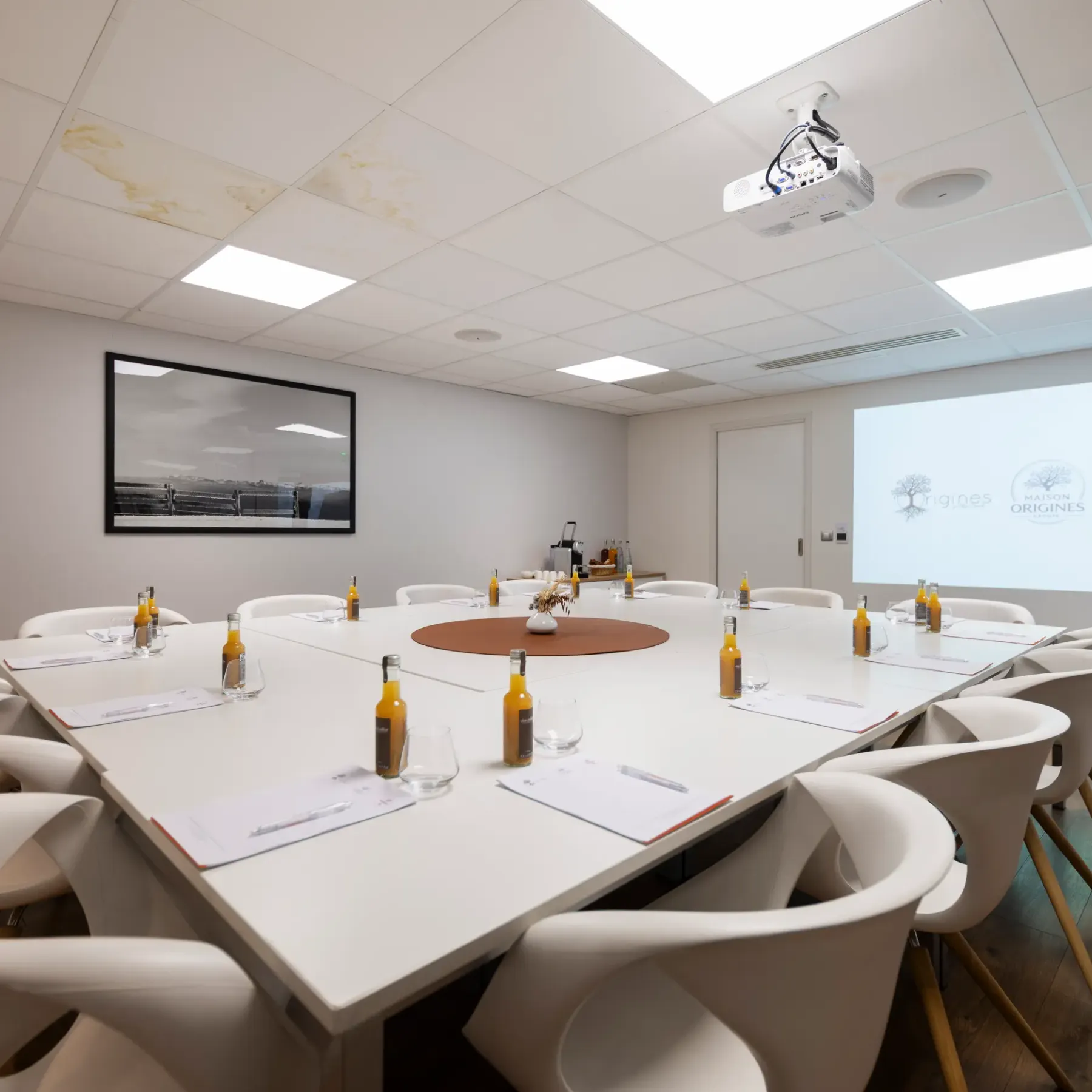 Salle de réunion avec une table ovale, des chaises modernes, des bouteilles et des verres, un projecteur et un écran affichant des logos