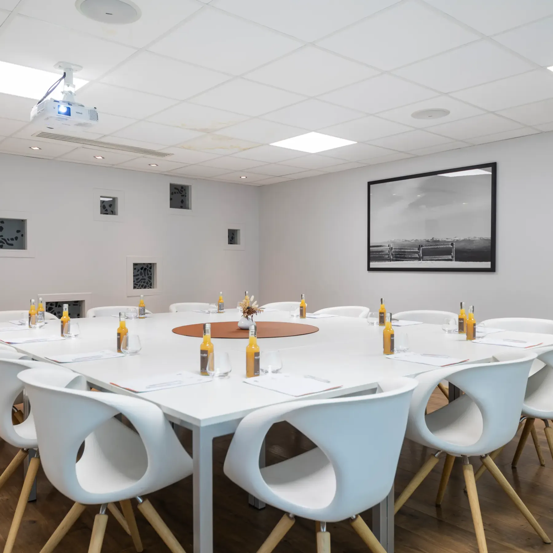 Salle de réunion avec une table ronde blanche entourée de chaises, des bouteilles et des verres posés sur la table, un projecteur au plafond et des œuvres d'art sur les murs