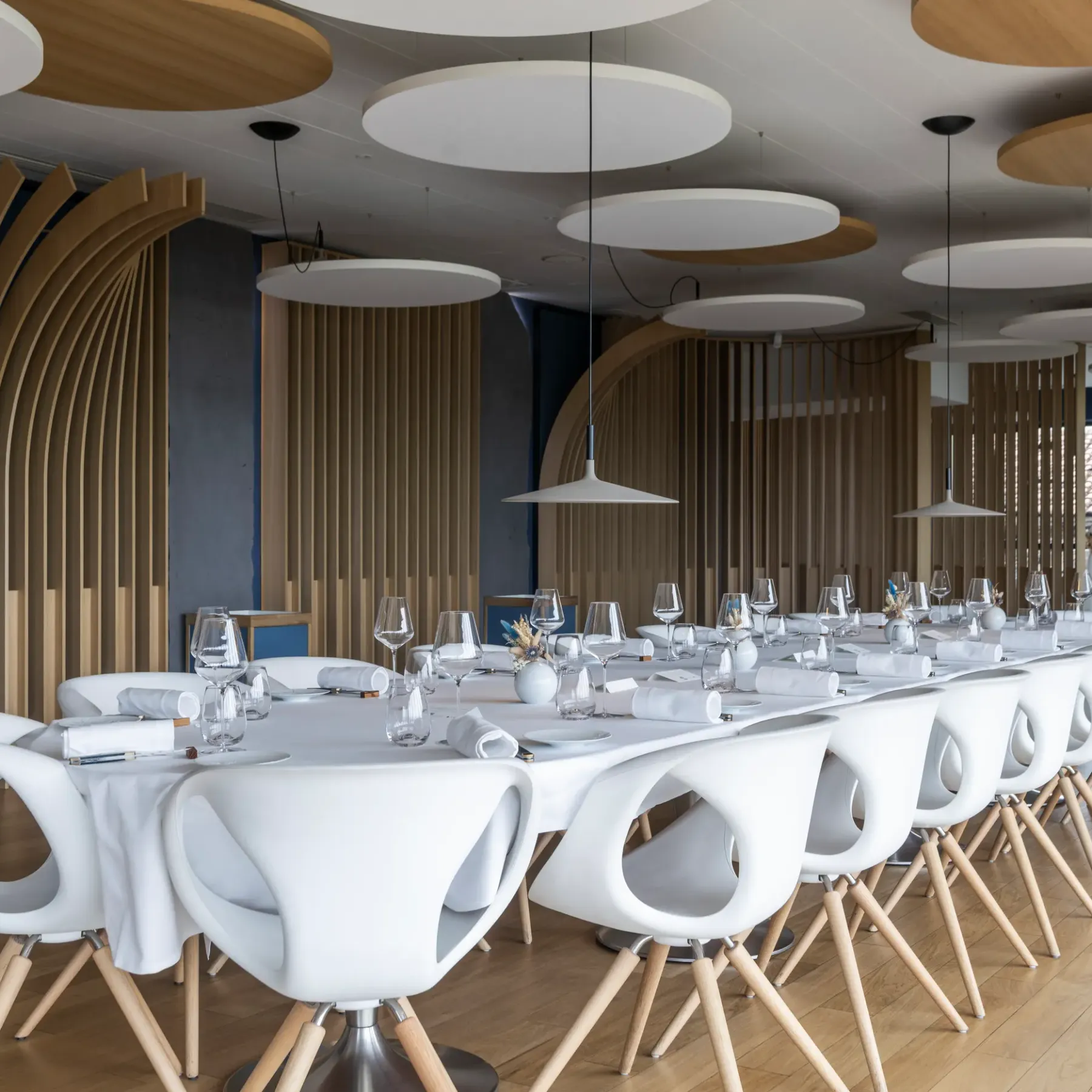 Une longue table rectangulaire avec des verres, des couverts et des serviettes, entourée de chaises blanches, dans une salle à manger moderne aux murs en bois et aux suspensions circulaires