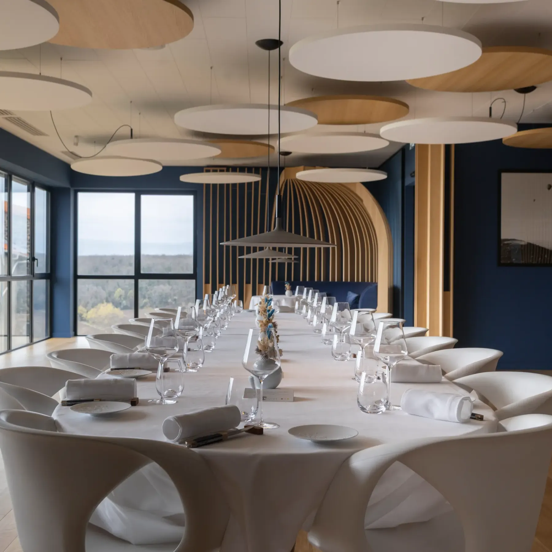 Une longue table de réunion avec des chaises modernes, des verres et des serviettes, dans une salle aux grandes fenêtres et aux luminaires circulaires suspendus