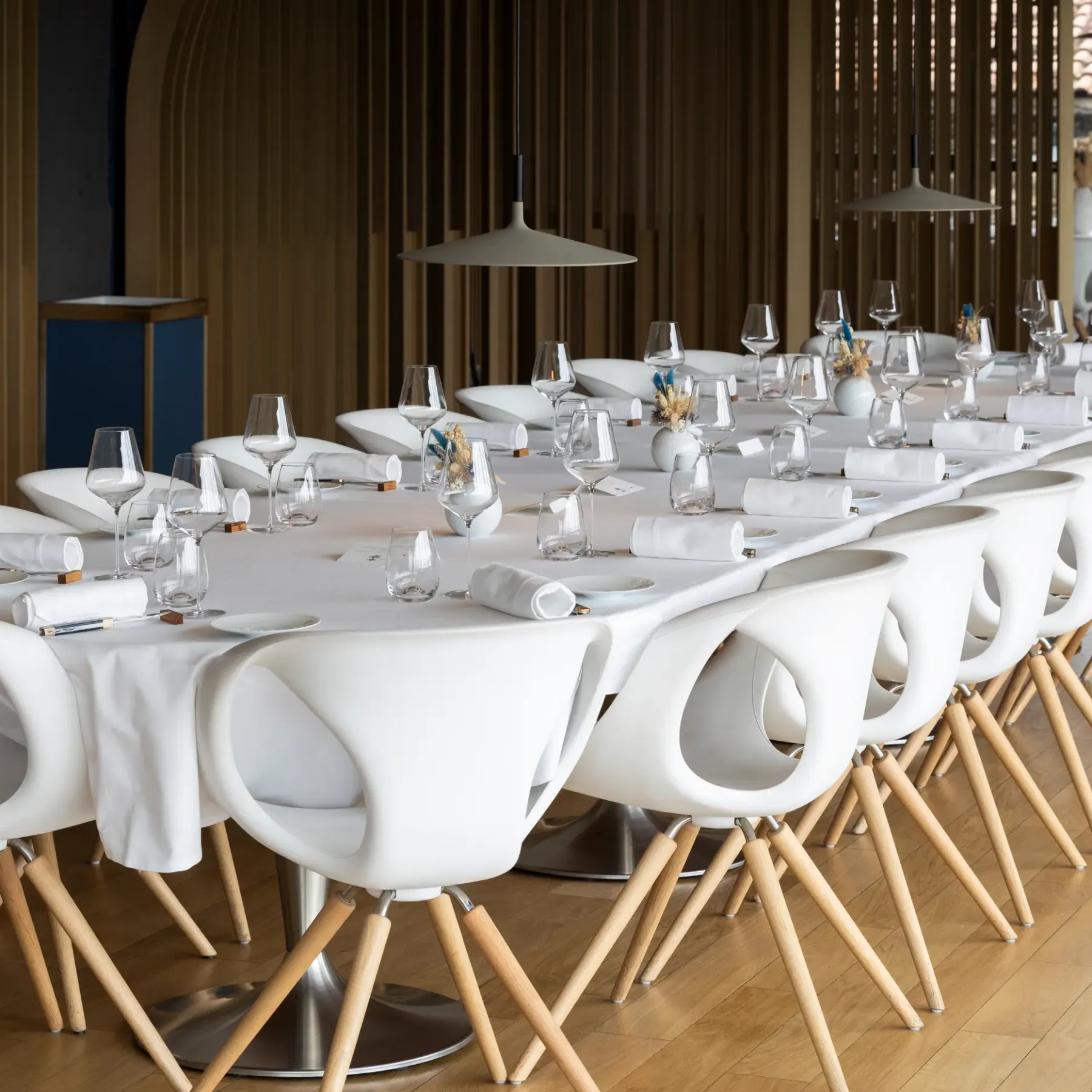 Une longue table rectangulaire est dressée avec des verres, des serviettes et des petits vases contenant des fleurs, entourée de chaises blanches aux pieds en bois, dans une salle à manger aux grandes fenêtres