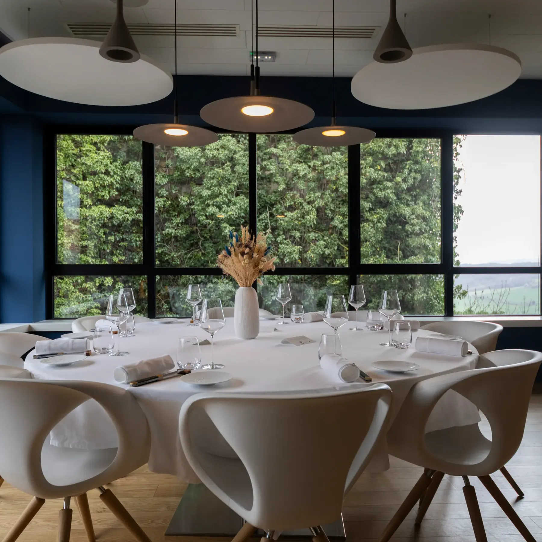 Une table ronde avec des verres et des couverts, entourée de chaises, dans une pièce avec de grandes fenêtres offrant une vue sur la nature