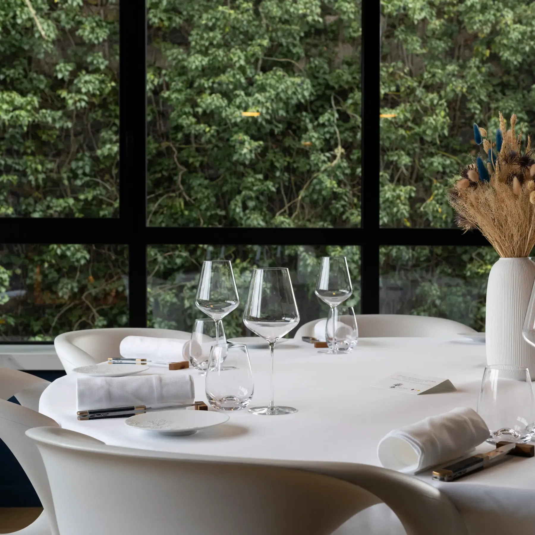 Table dressée avec verres à vin, serviettes et bouquet de fleurs séchées devant une grande fenêtre donnant sur une forêt
