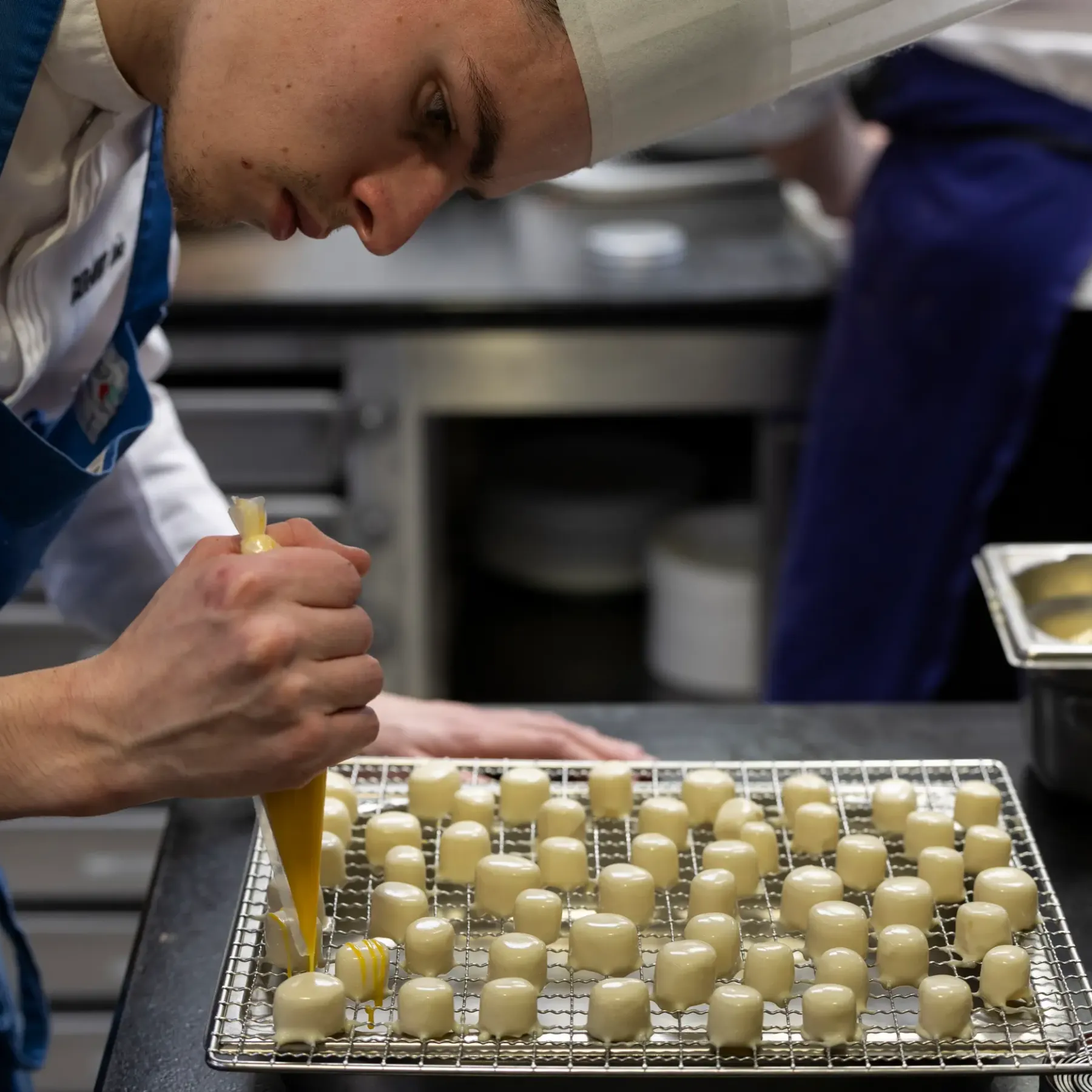 Un cuisinier en uniforme verse une préparation jaune sur des petits carrés de pâte disposés sur une grille métallique