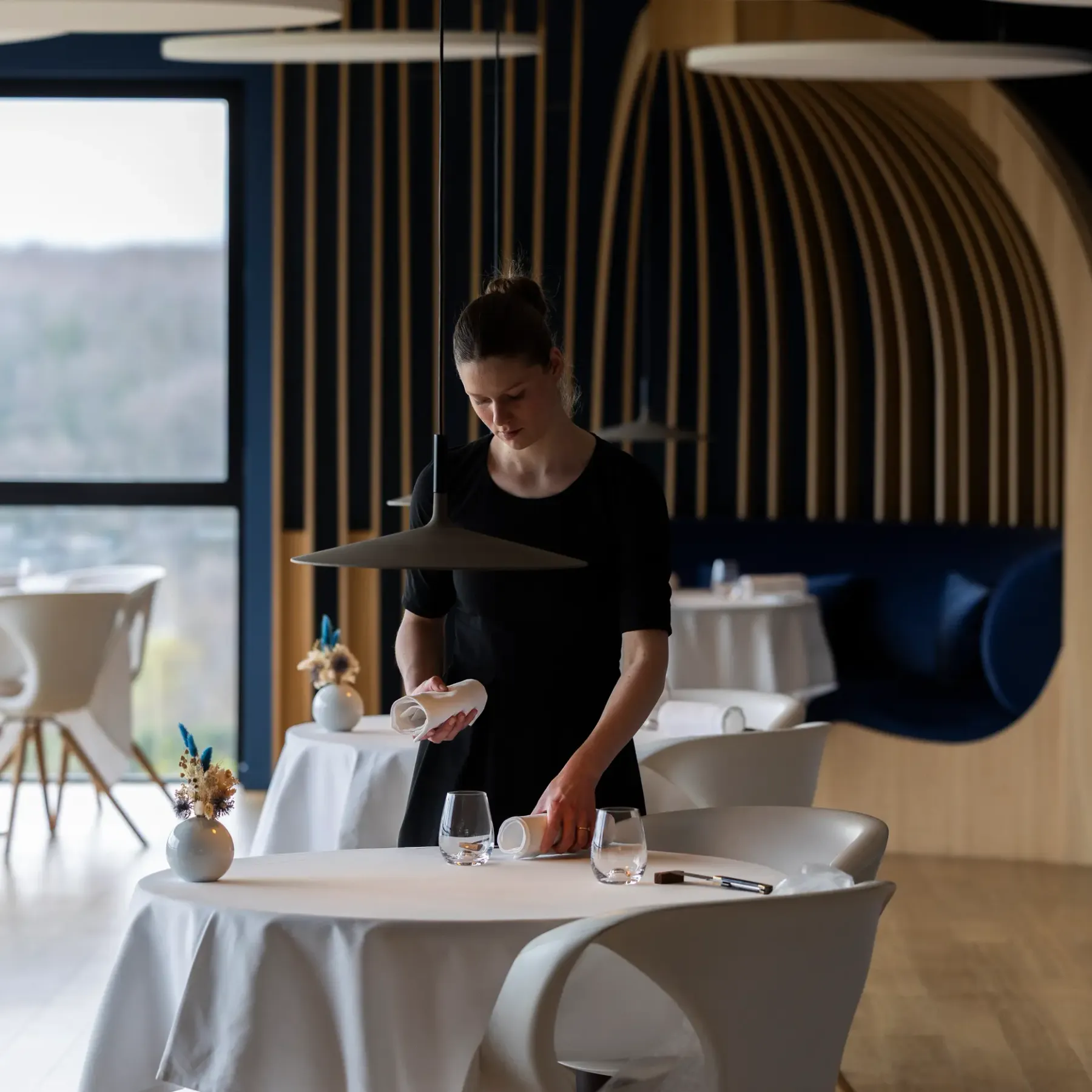 Une personne en tenue noire prépare une table dans un restaurant moderne aux grandes baies vitrées