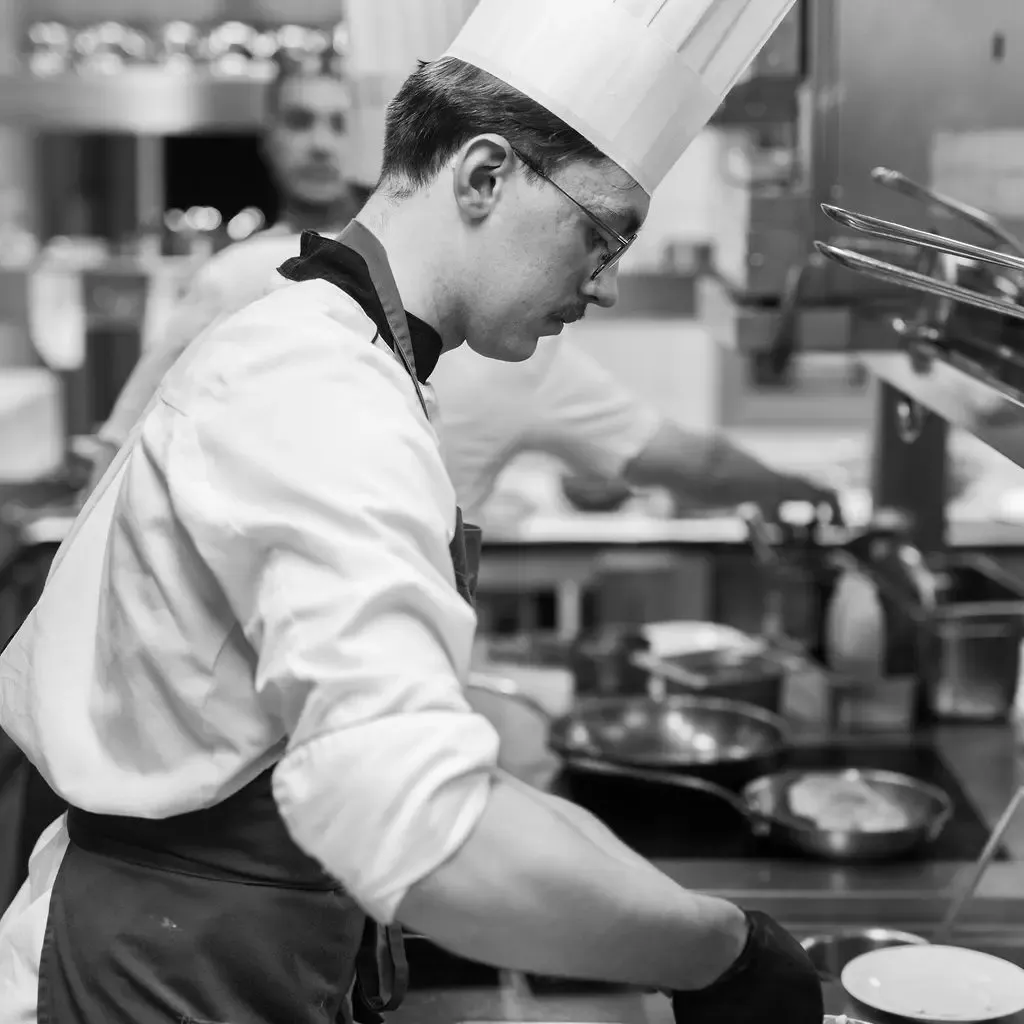 Un chef cuisinier en toque blanche travaille dans une cuisine professionnelle, concentré sur sa préparation culinaire