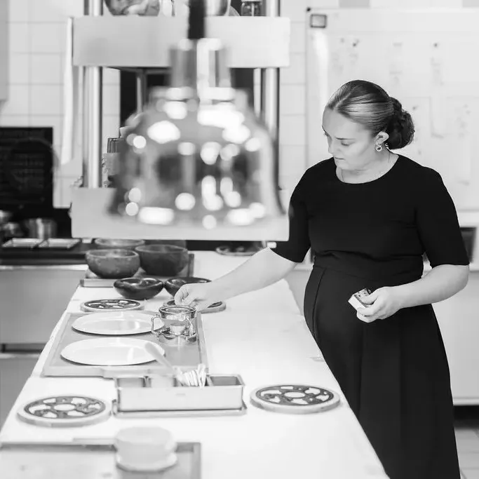 A woman in a black dress prepares plates in a professional kitchen equipped with various utensils and containers.