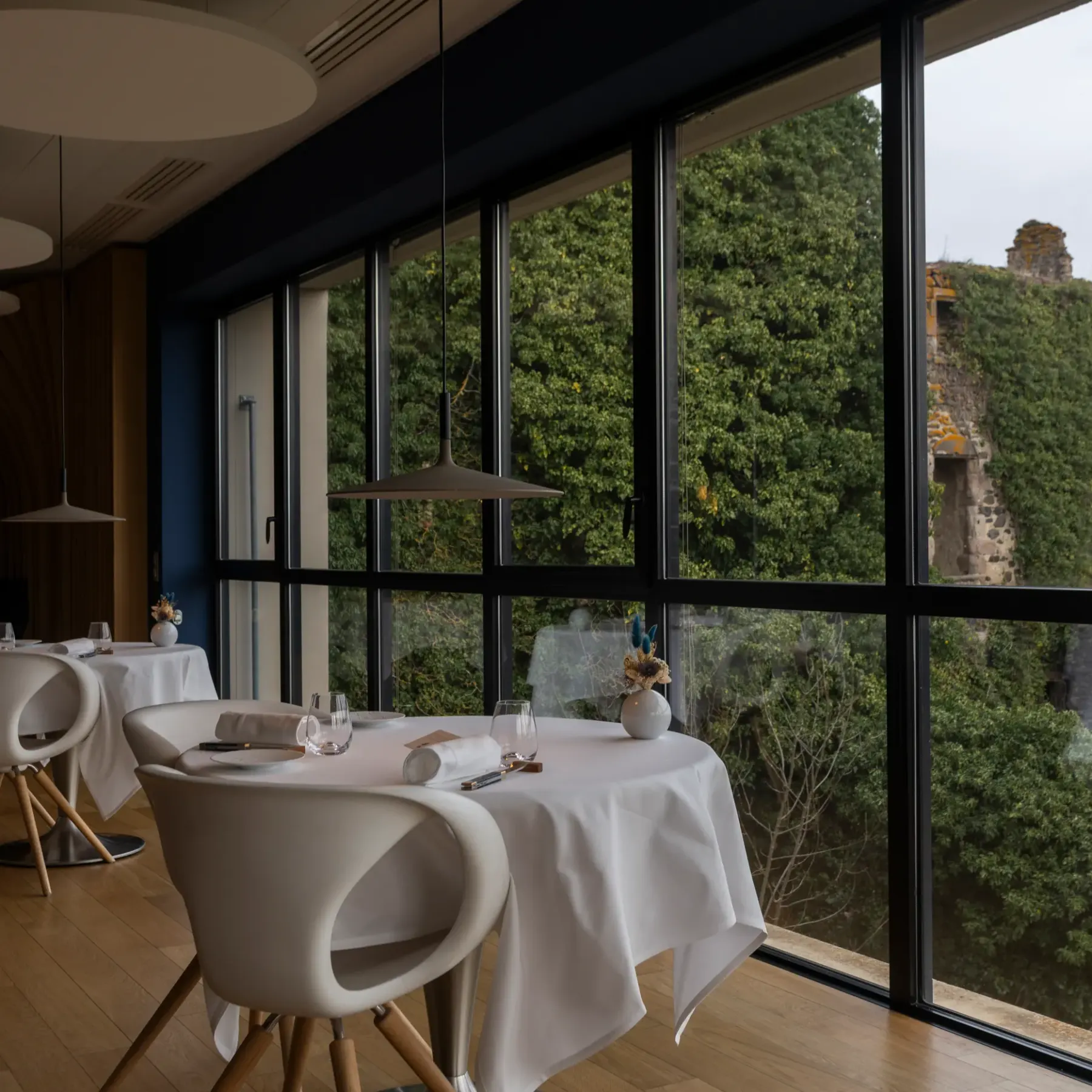 Restaurant avec tables et chaises blanches devant de grandes baies vitrées offrant une vue sur une colline boisée et des ruines