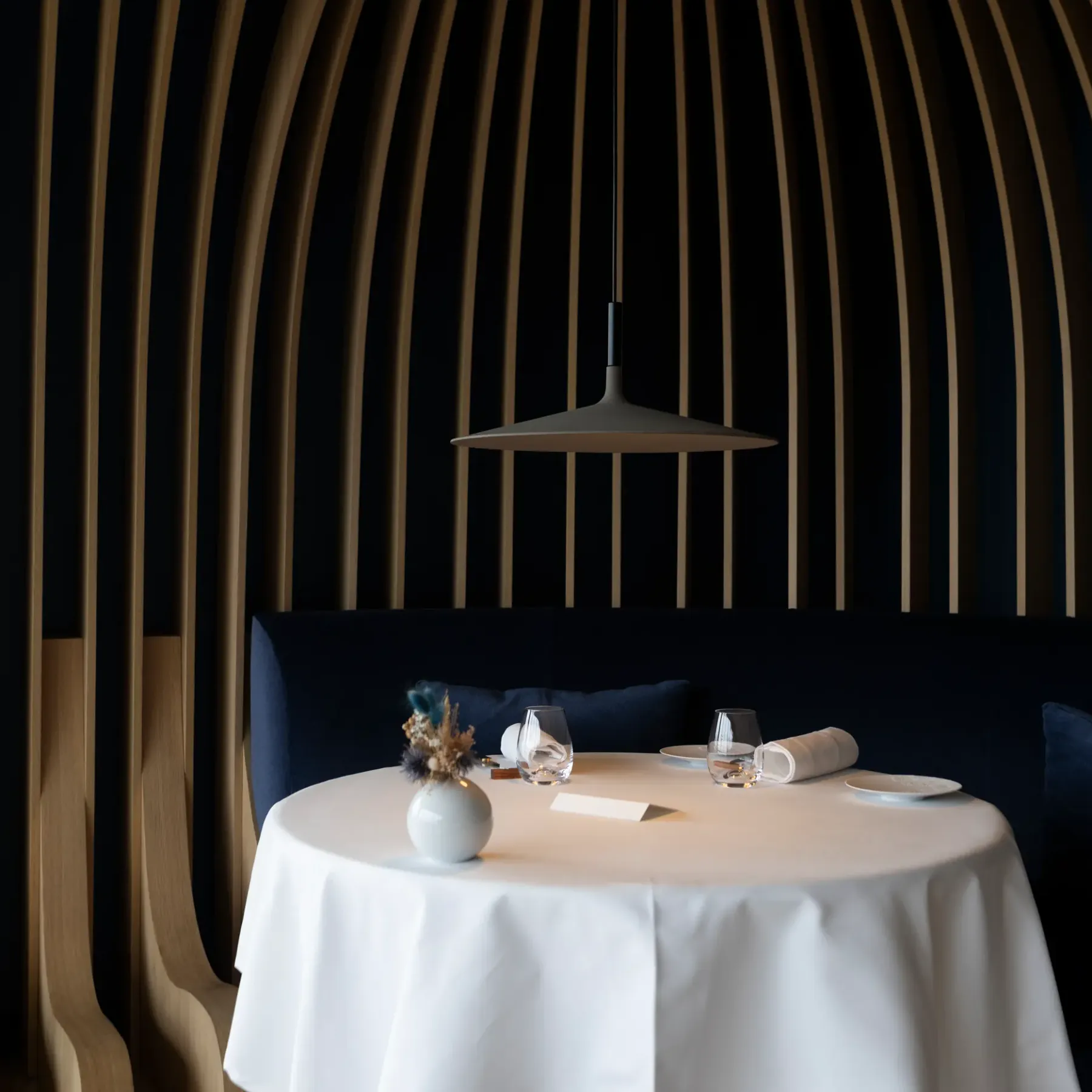 Table dressée avec nappe blanche, vase de fleurs, verres et serviette roulée, sous une suspension, dans un espace aux murs à rayures verticales sombres et claires