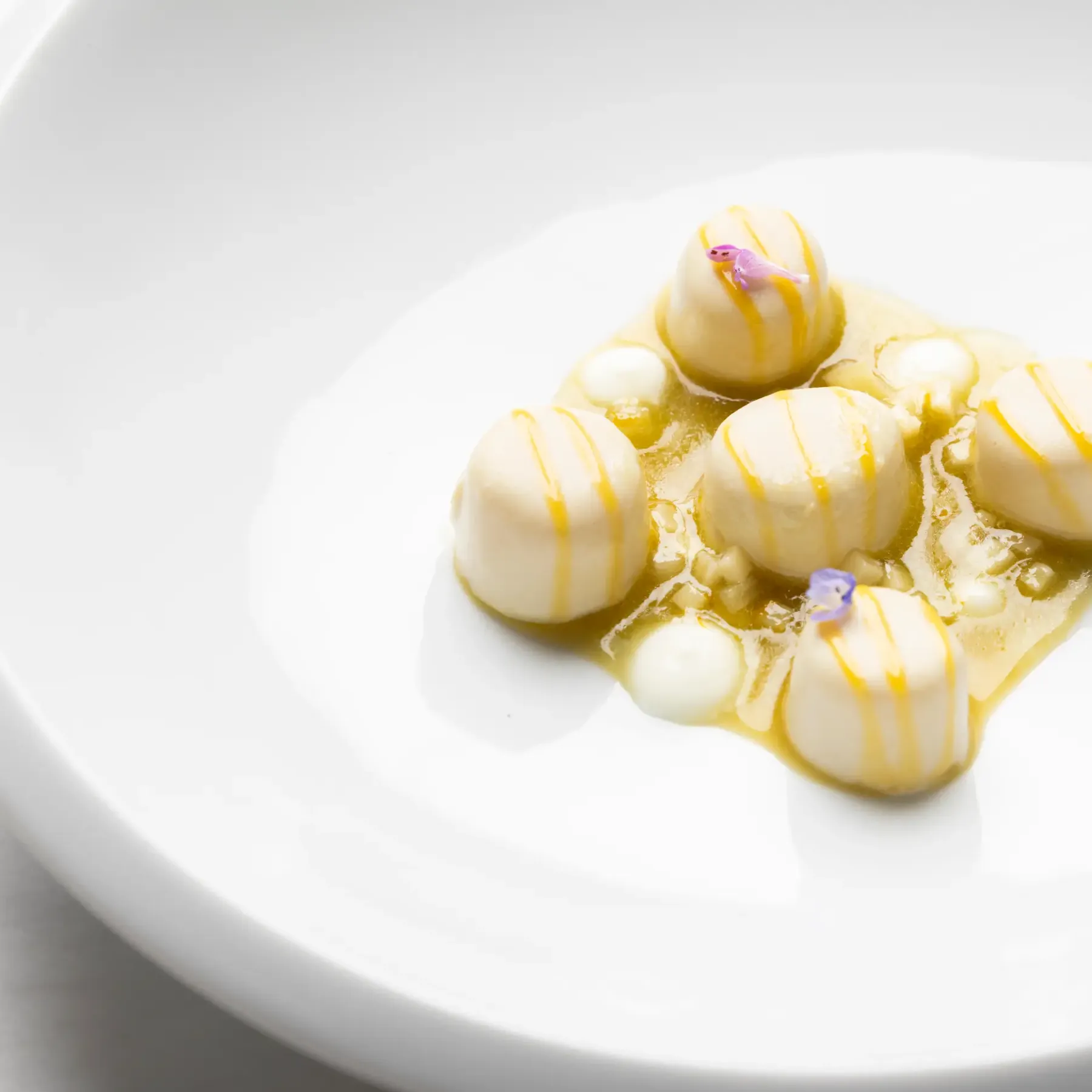 Des sphères de dessert blanc sur une sauce jaune clair, décorées de petites fleurs violettes, présentées sur une assiette blanche