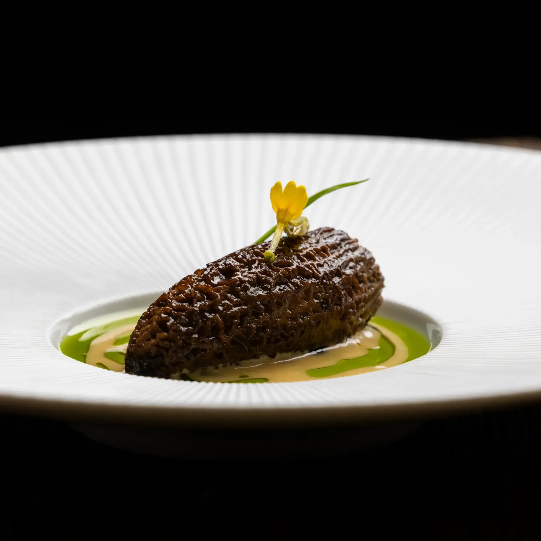 Un morceau de nourriture brun foncé est placé sur une assiette blanche avec une sauce verte autour, accompagné d'une petite fleur jaune