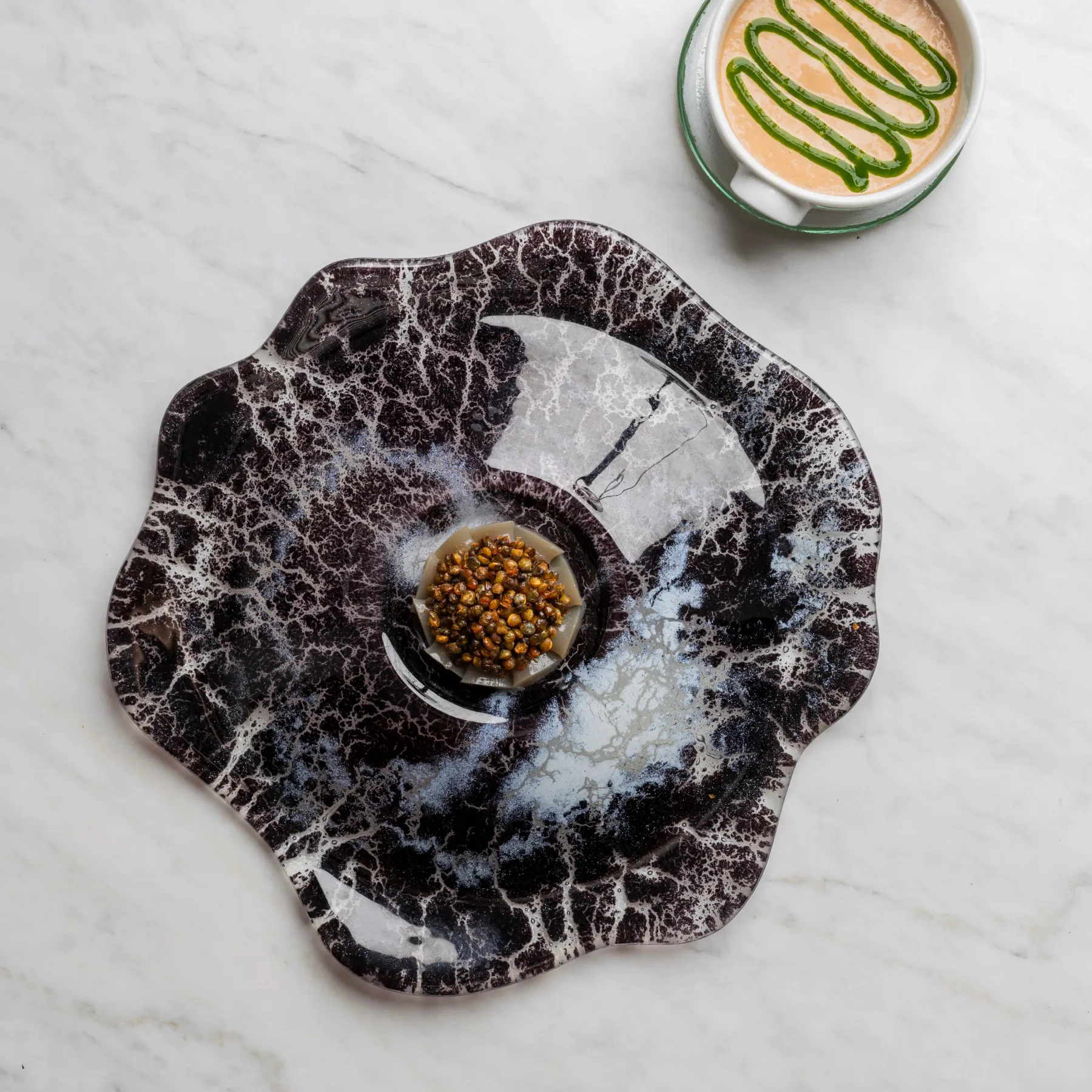 Une tasse de café avec une décoration verte à la surface est posée sur une soucoupe, accompagnée d'une assiette contenant des graines sur une surface en marbre blanc