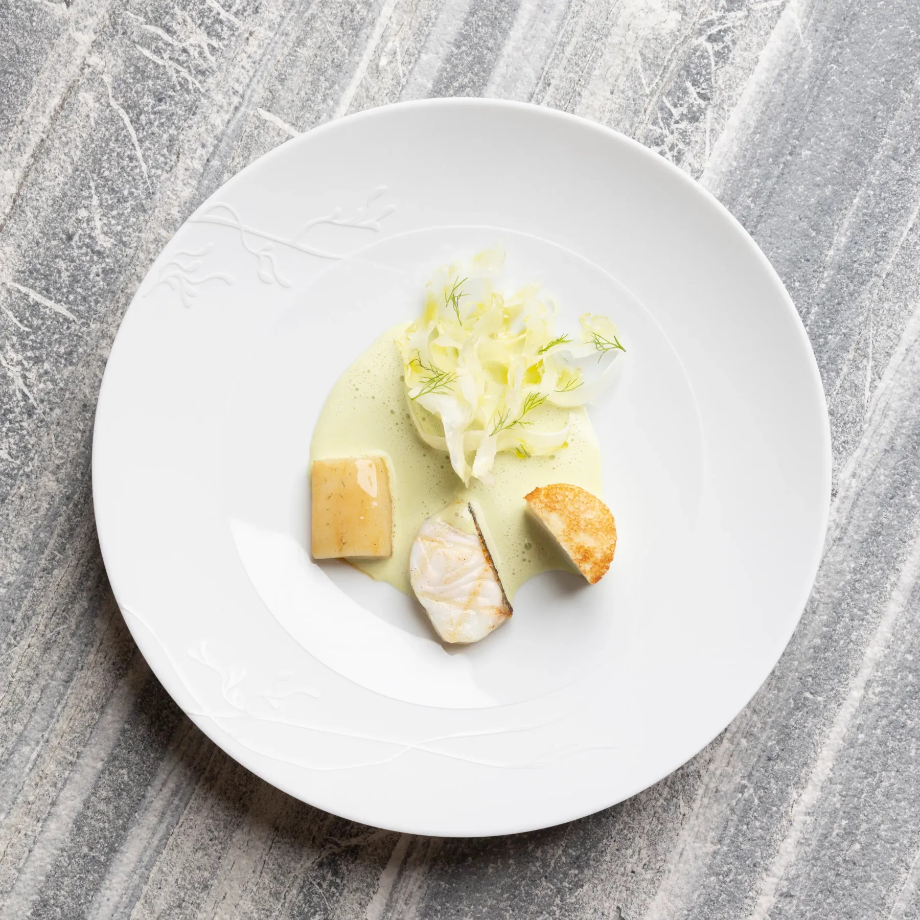 Un plat avec une sauce verte, des morceaux de poisson, des légumes et des croûtons sur une assiette blanche posée sur une surface en marbre gris