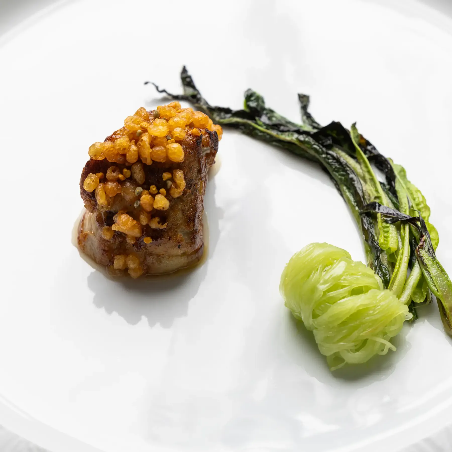 Un morceau de viande garni de grains dorés accompagné de légumes verts et d'une boule translucide sur une assiette blanche