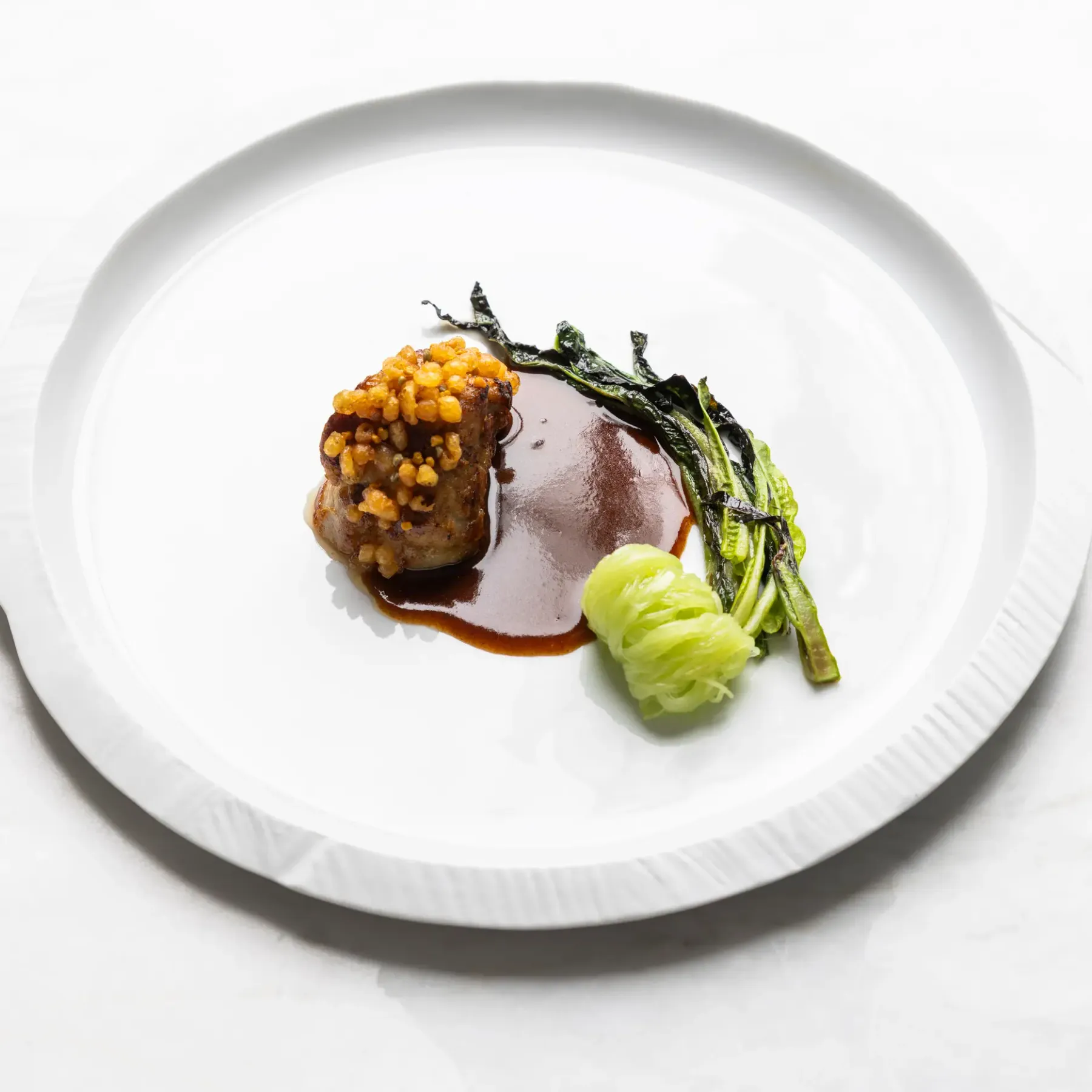 Un plat composé d'un morceau de viande recouvert de graines, accompagné de légumes verts et d'une sauce, présenté sur une assiette blanche