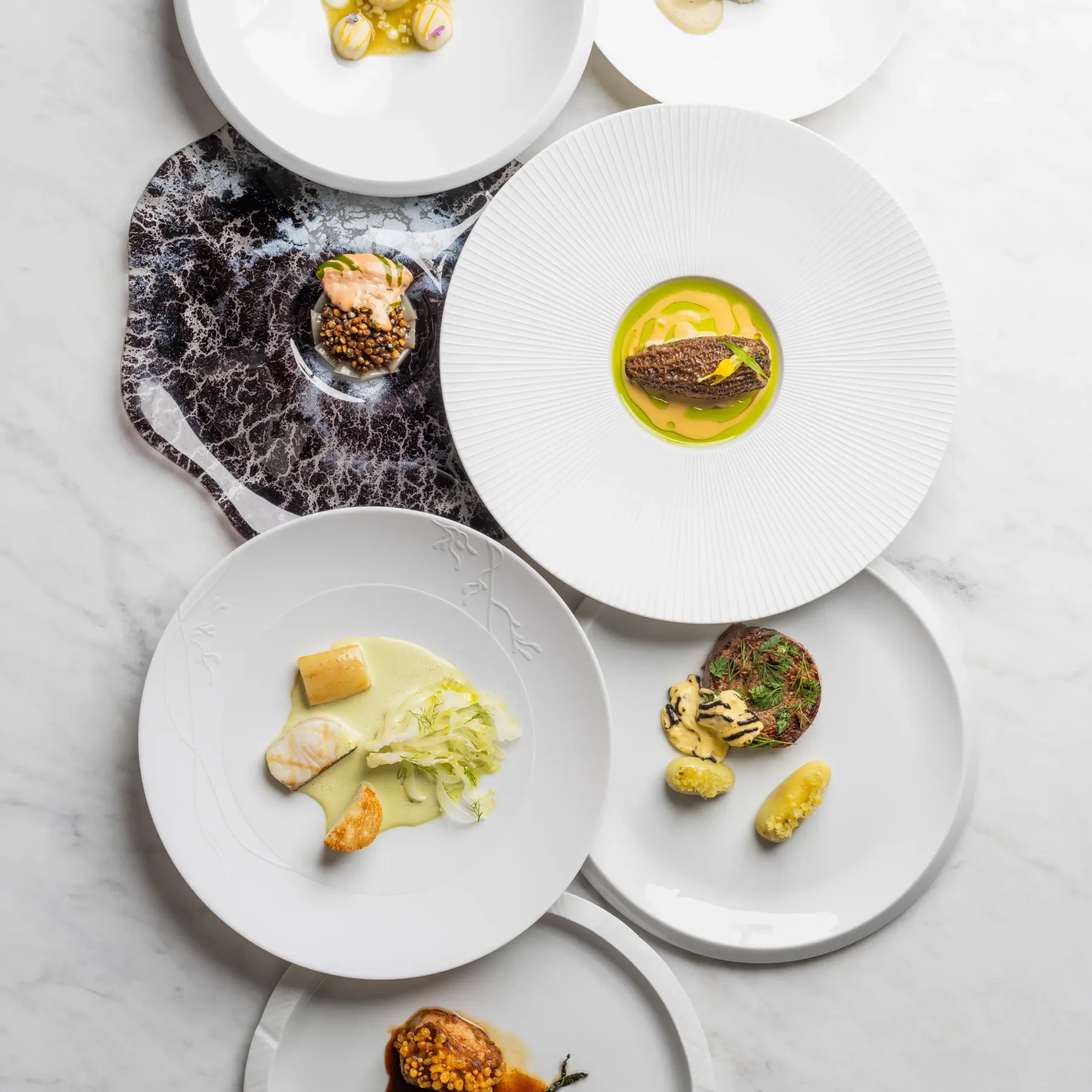Six assiettes blanches présentant des plats gastronomiques variés disposés sur une surface en marbre