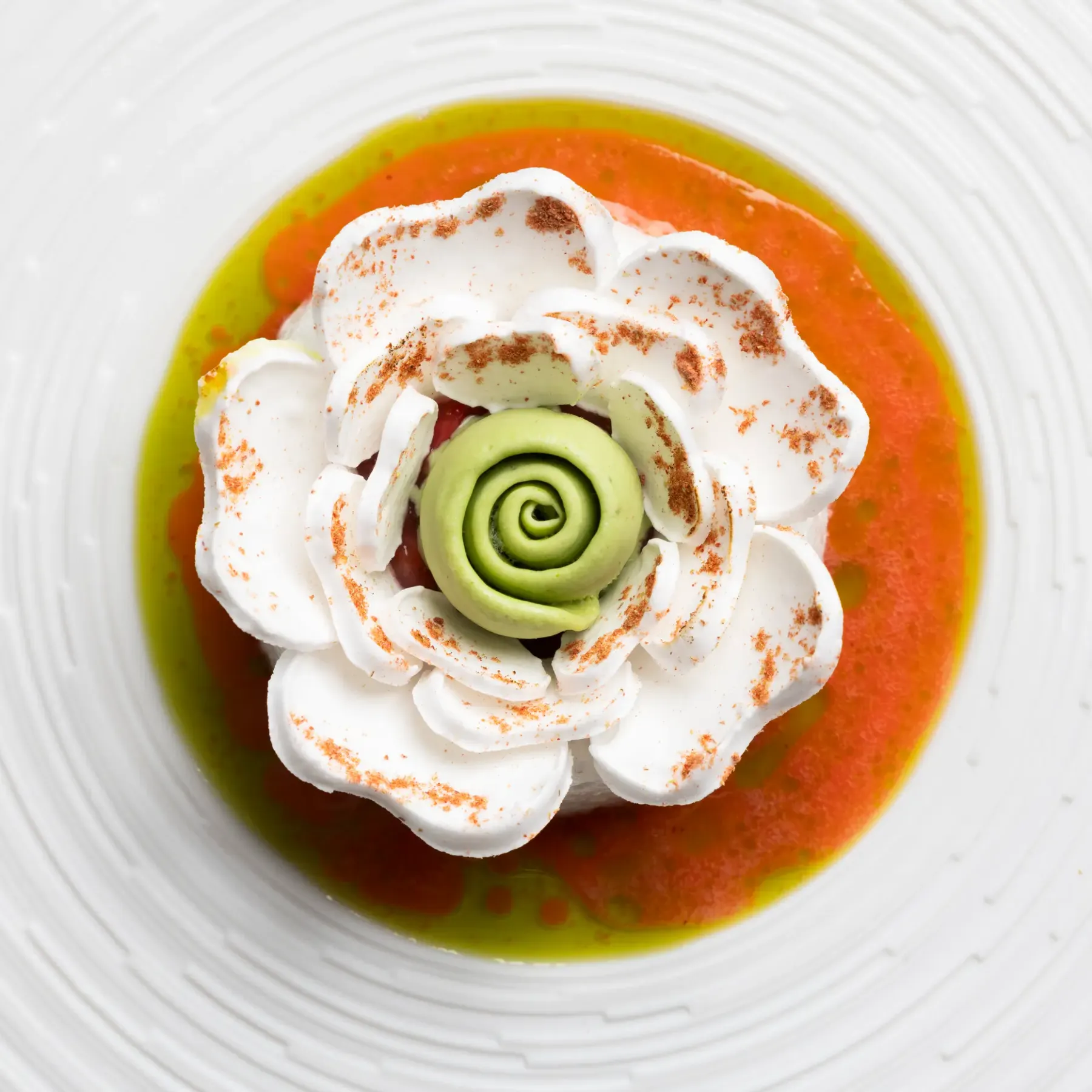 Un dessert composé d'une mousse blanche en forme de fleur, d'une spirale verte et d'une sauce orange et verte dans une assiette blanche