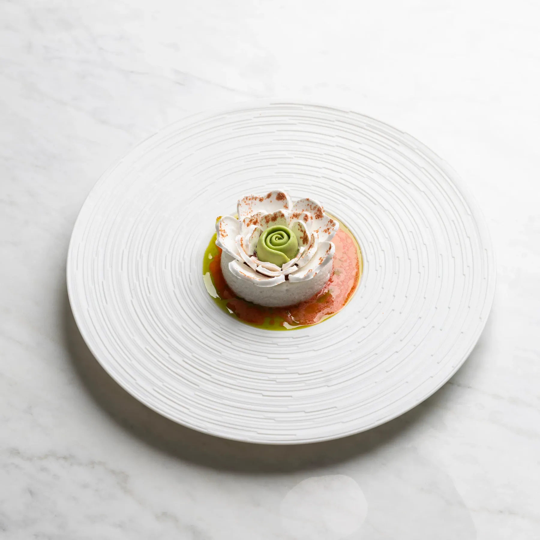 Un petit dessert en forme de rose blanche avec une touche verte au centre, posé sur une assiette blanche circulaire sur une surface en marbre blanc
