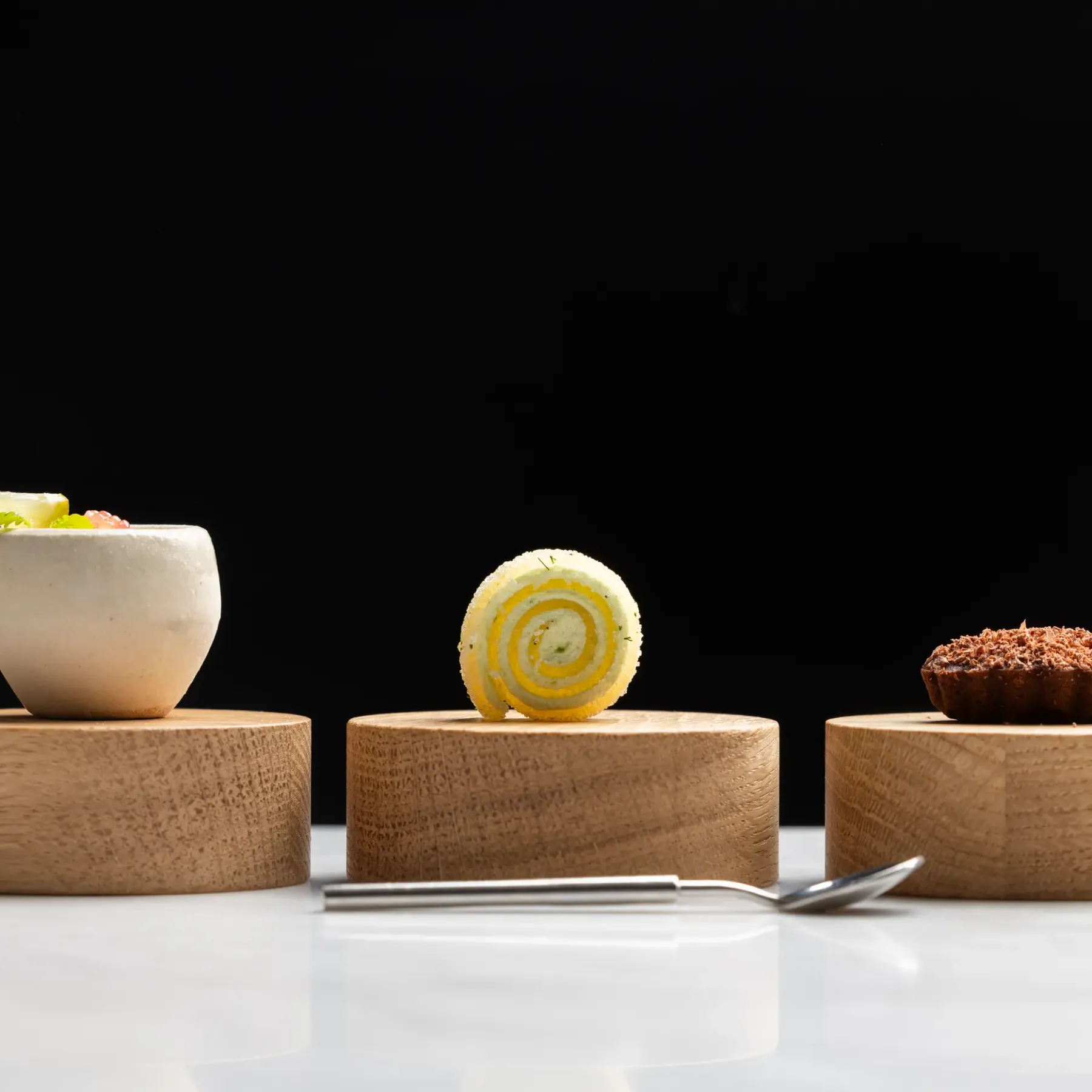 Trois desserts présentés sur des socles en bois clair, dont une coupe blanche avec garniture, une tranche enroulée et un petit gâteau recouvert de cacao